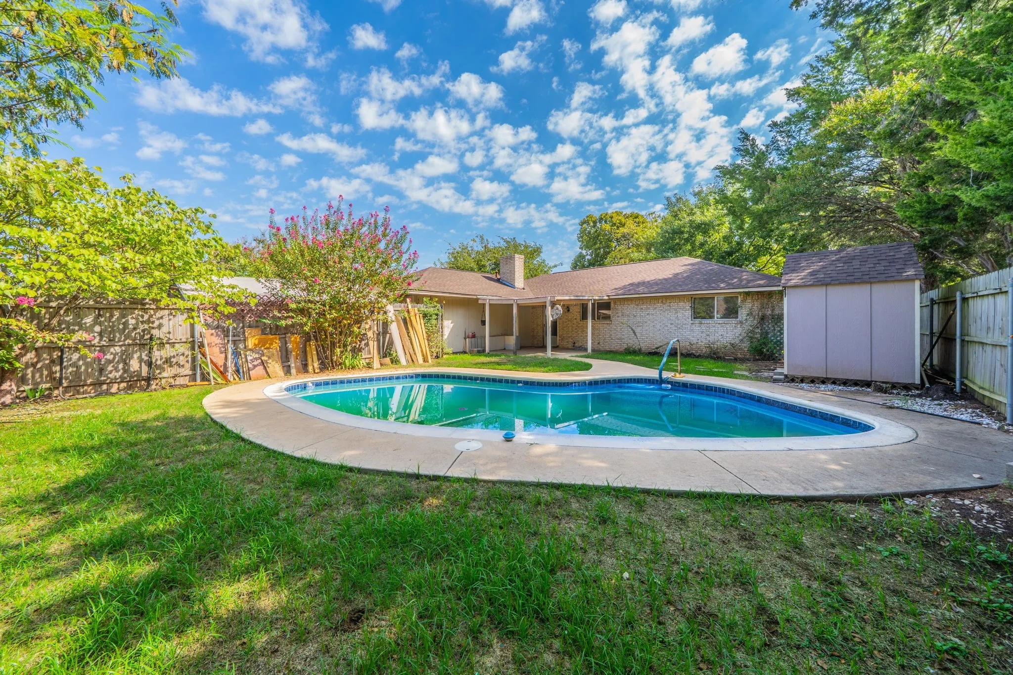 Backyard featuring a kidney-shaped swimming pool with concrete decking, surrounding grassy areas, and a wooden privacy fence