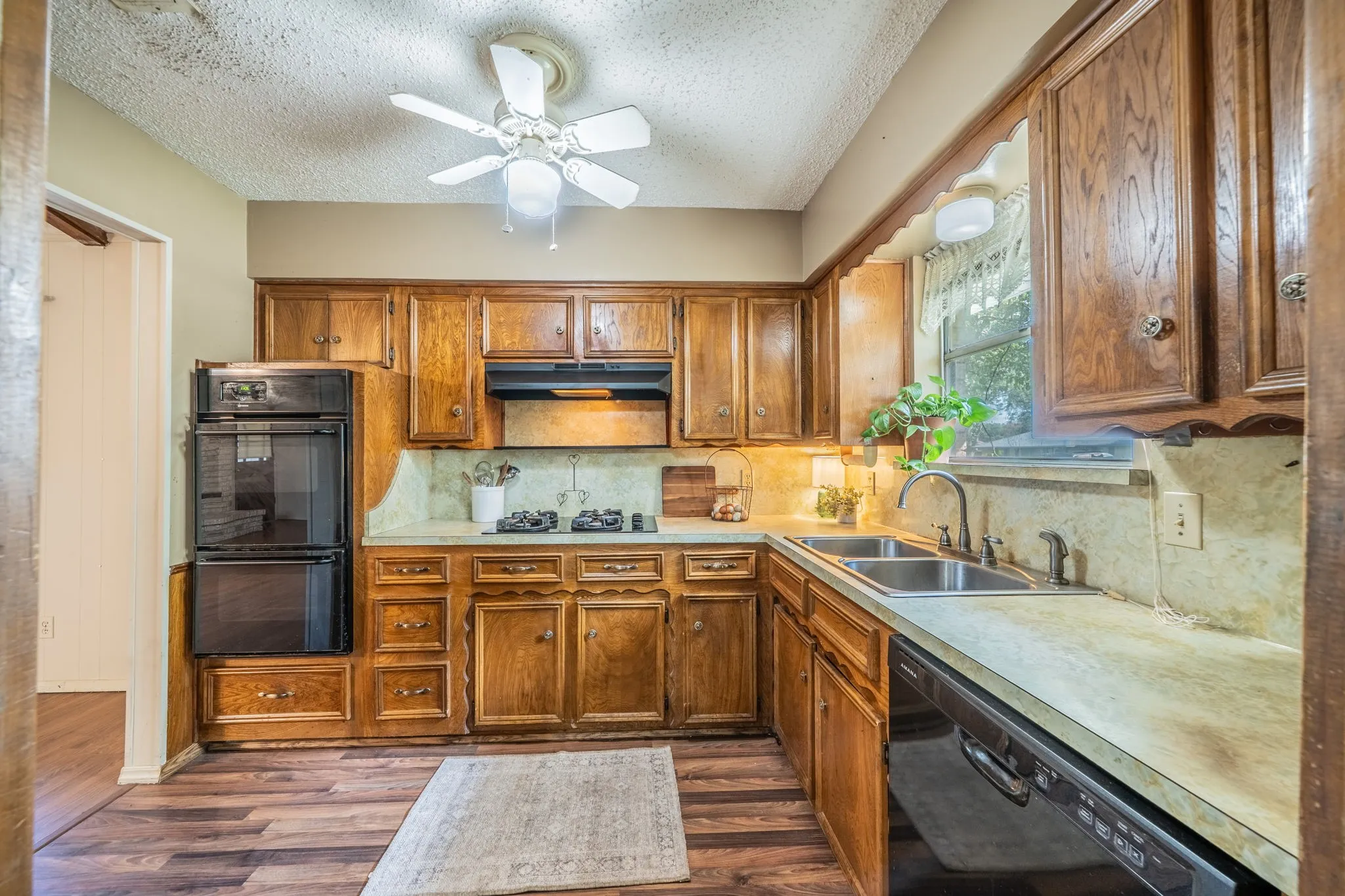 Wood-finish flooring, wood cabinetry, built-in double ovens, dual basin sink, and a ceiling fan with integrated lighting