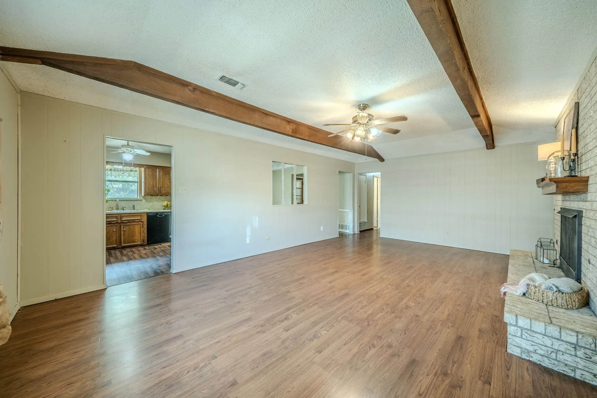 Spacious living area featuring wood-finish flooring, exposed ceiling beams, a ceiling fan, and a brick fireplace with a wood mantle