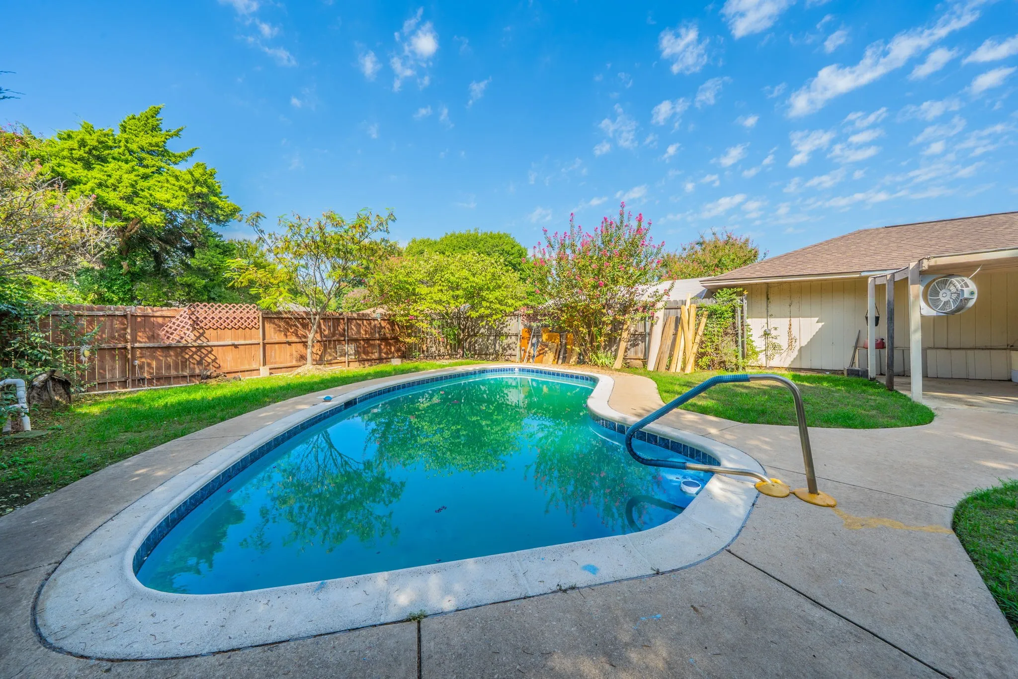 Curved swimming pool with a concrete surround, wood privacy fencing, and green lawn areas