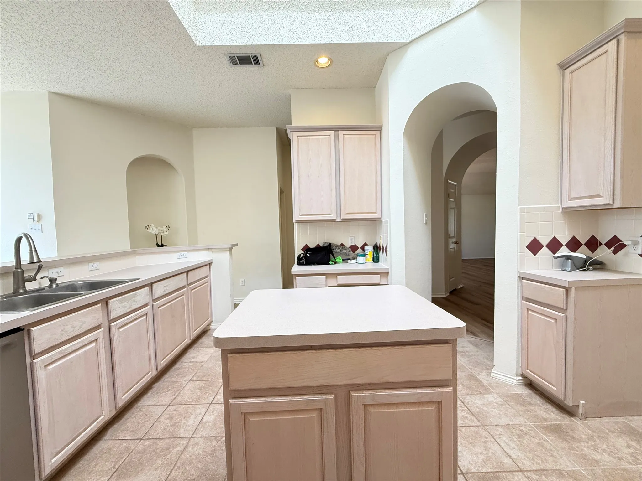 Kitchen featuring tile flooring, light wood-finish cabinetry, a stainless steel sink, a central island, and arched doorways