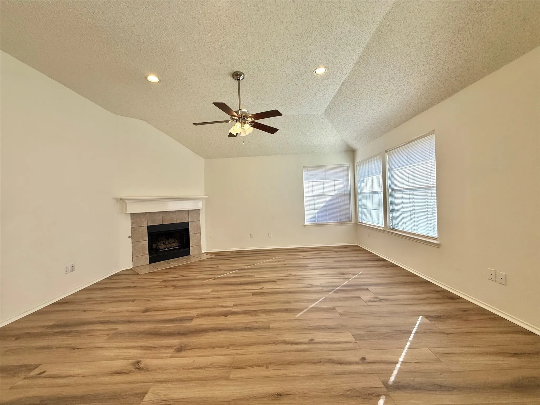 Vaulted ceiling, wood-finish flooring, corner fireplace with tile surround, multiple windows, and a ceiling fan with integrated lighting