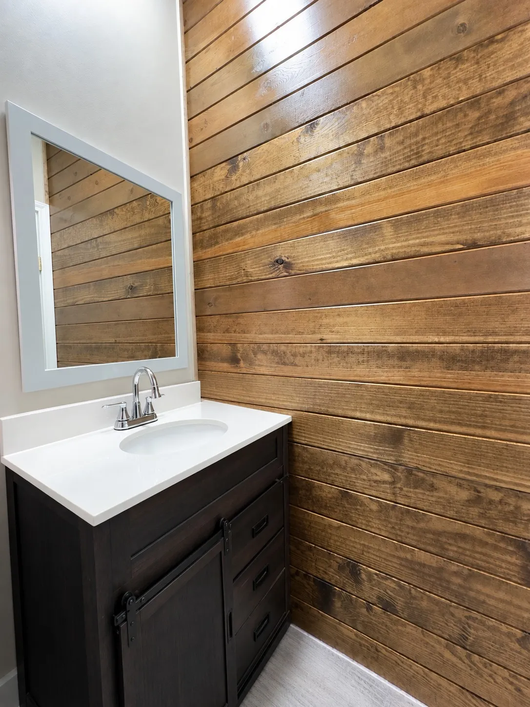 Guest. Powder room with a wood plank accent wall, dark wood-finish vanity with barn door hardware, white countertop, undermount sink, and chrome faucet