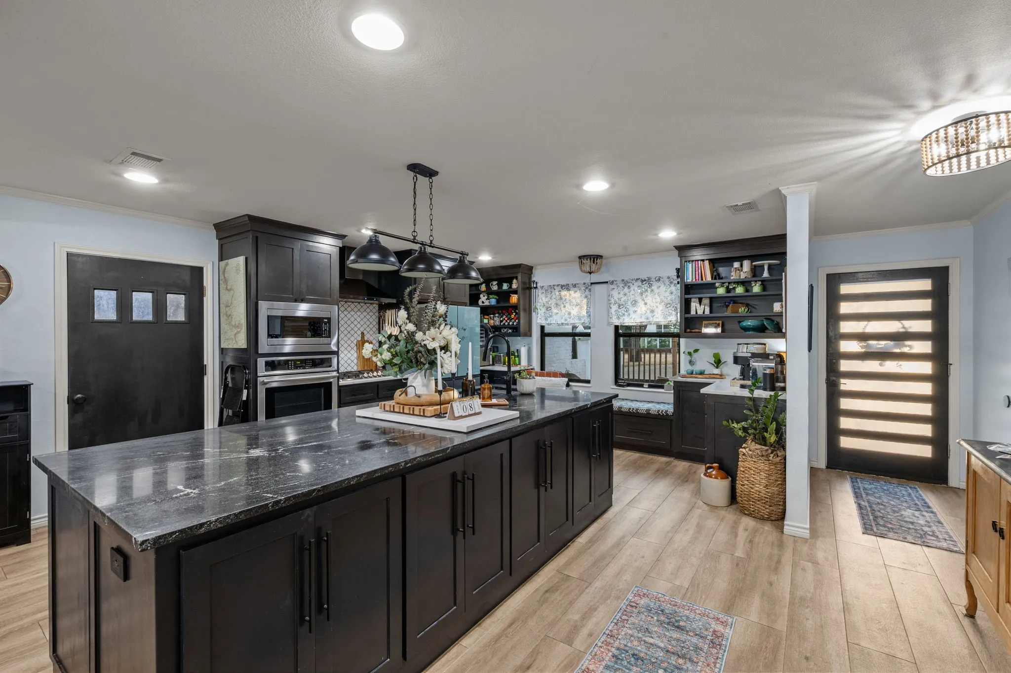 Expansive kitchen island with a dark stone countertop, dark cabinetry, stainless steel appliances, wood-finish flooring, and built-in shelving