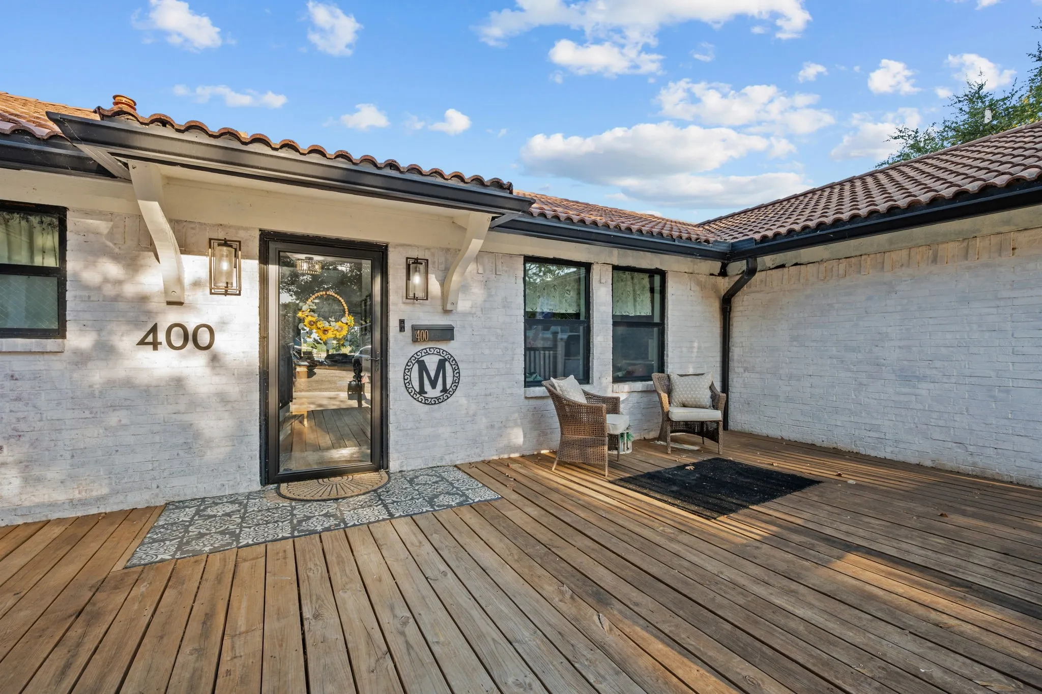 Expansive wood-finish decking, white brick exterior, black frame windows, ceramic tile roofing, and modern exterior lighting fixtures