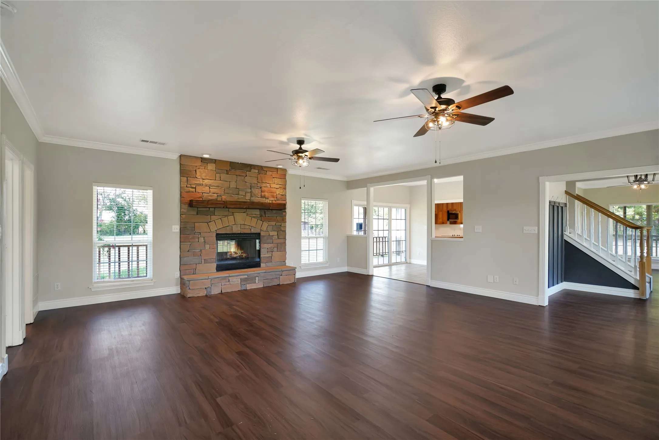 Spacious living area featuring wood-finish flooring, a stone-clad fireplace with a wood mantel, multiple windows, and ceiling fans