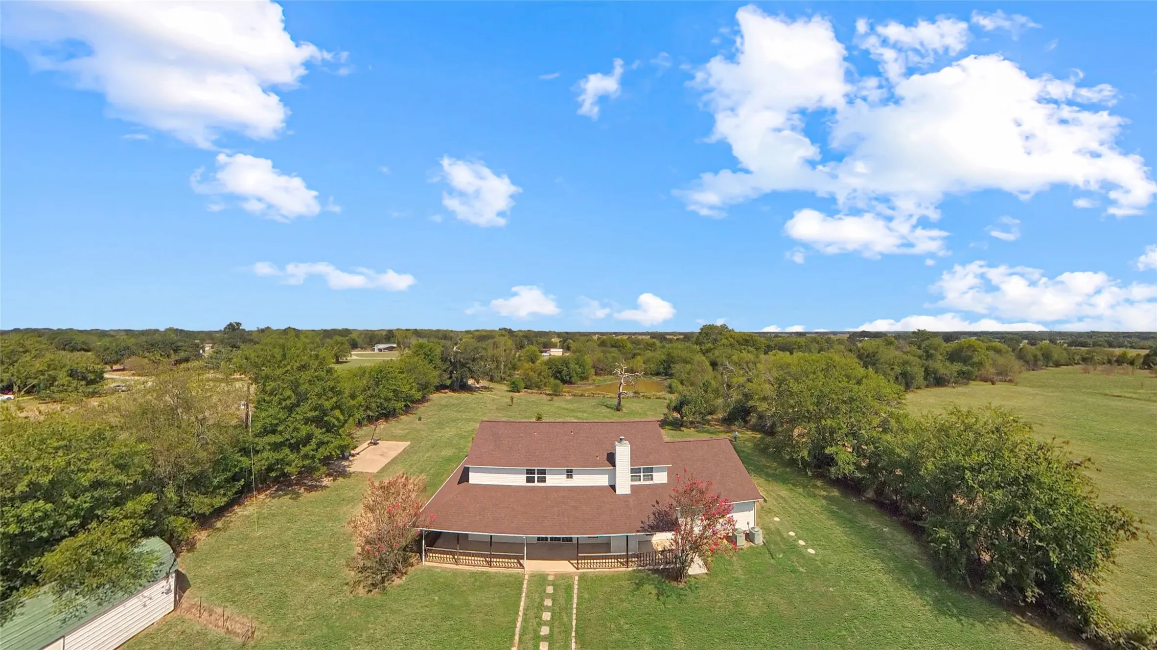 Expansive property featuring a residence with a brown shingle roof and white siding, surrounded by mature trees, and a substantial green lawn