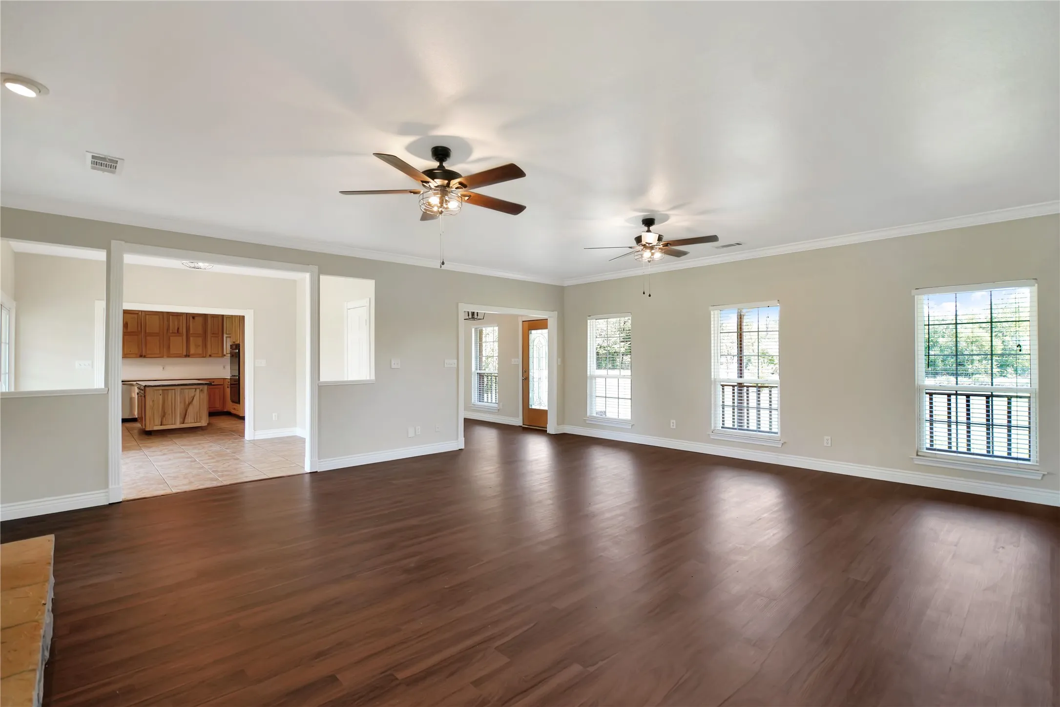 Spacious living area featuring wood-finish flooring, two ceiling fans with integrated lighting, multiple windows, and crown molding