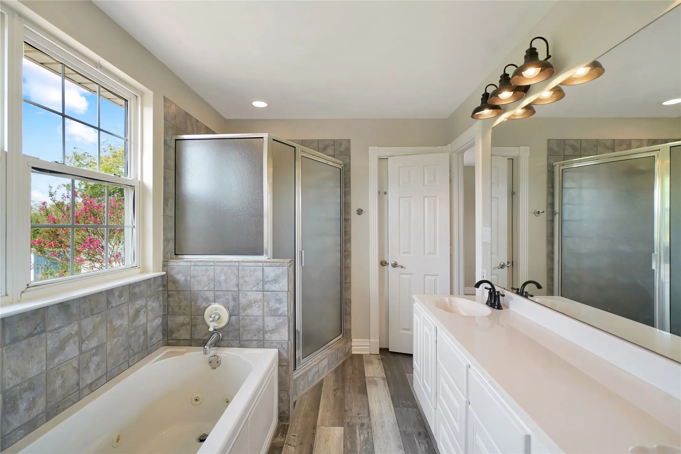 Dual-sink vanity with a light-toned countertop, mirrored wall, wood-finish flooring, a jetted tub, and a separate shower enclosure