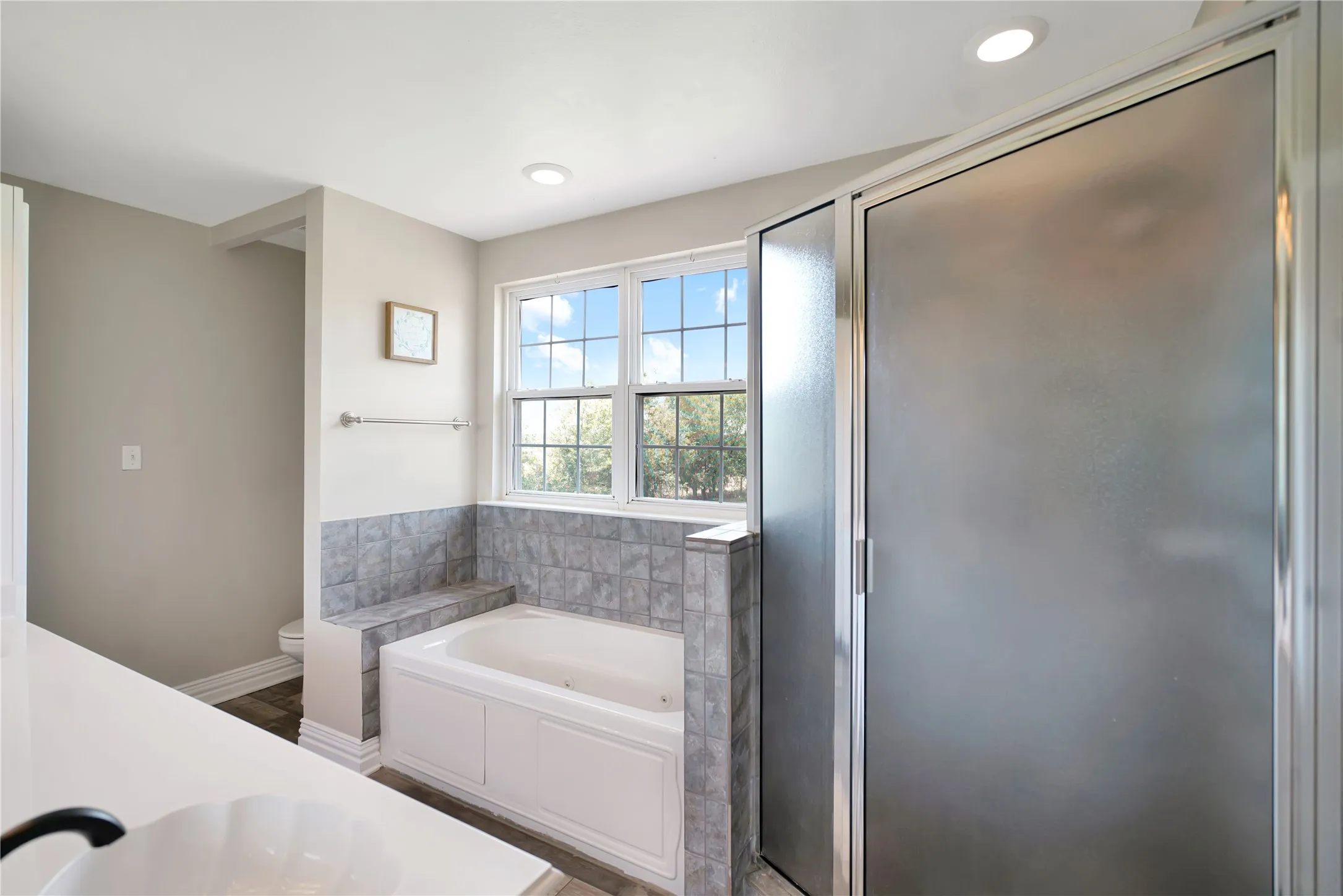 Built-in soaking tub with tile surround, glass-enclosed shower, dual-pane window, and recessed lighting