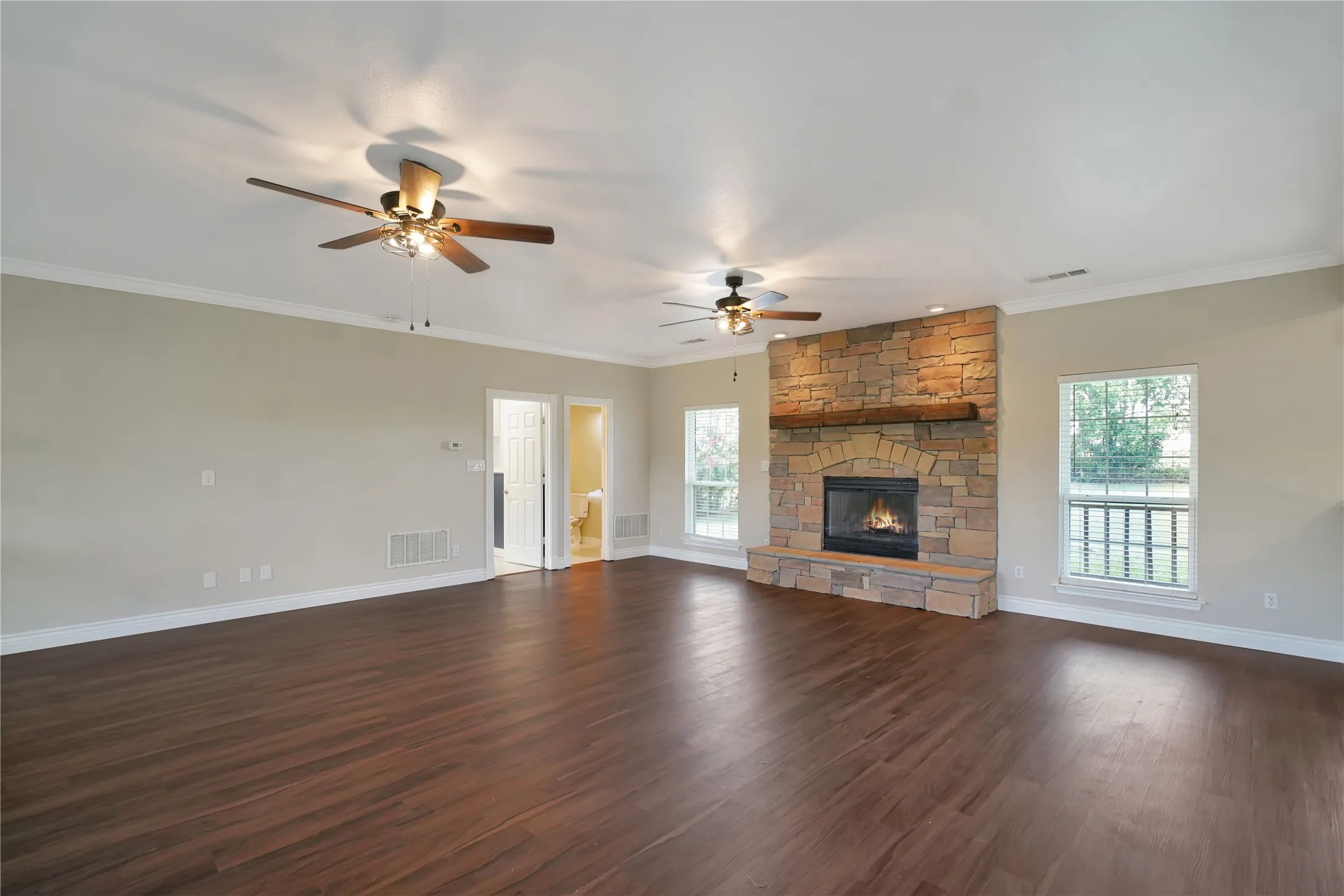 Spacious living area featuring wood-finish flooring, a stone-clad fireplace with a wood mantel, multiple windows, and two ceiling fans with integrated lighting