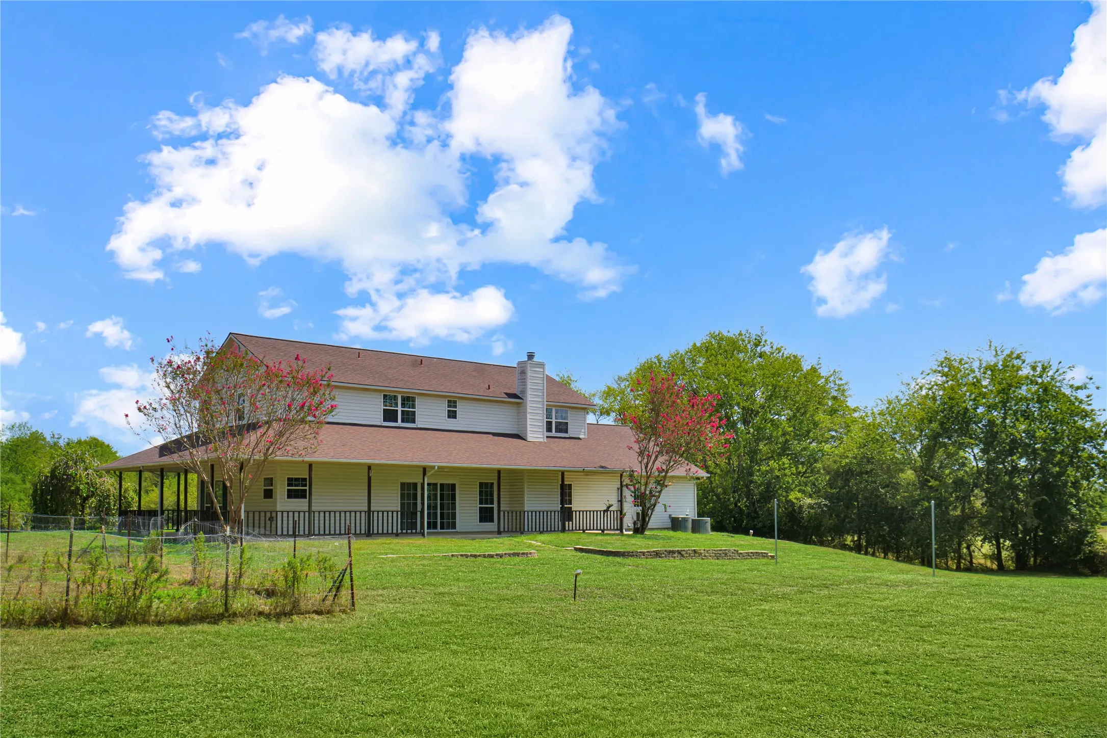 Expansive grassy yard with mature trees, a two-story residence featuring a brown shingle roof, and a perimeter fence