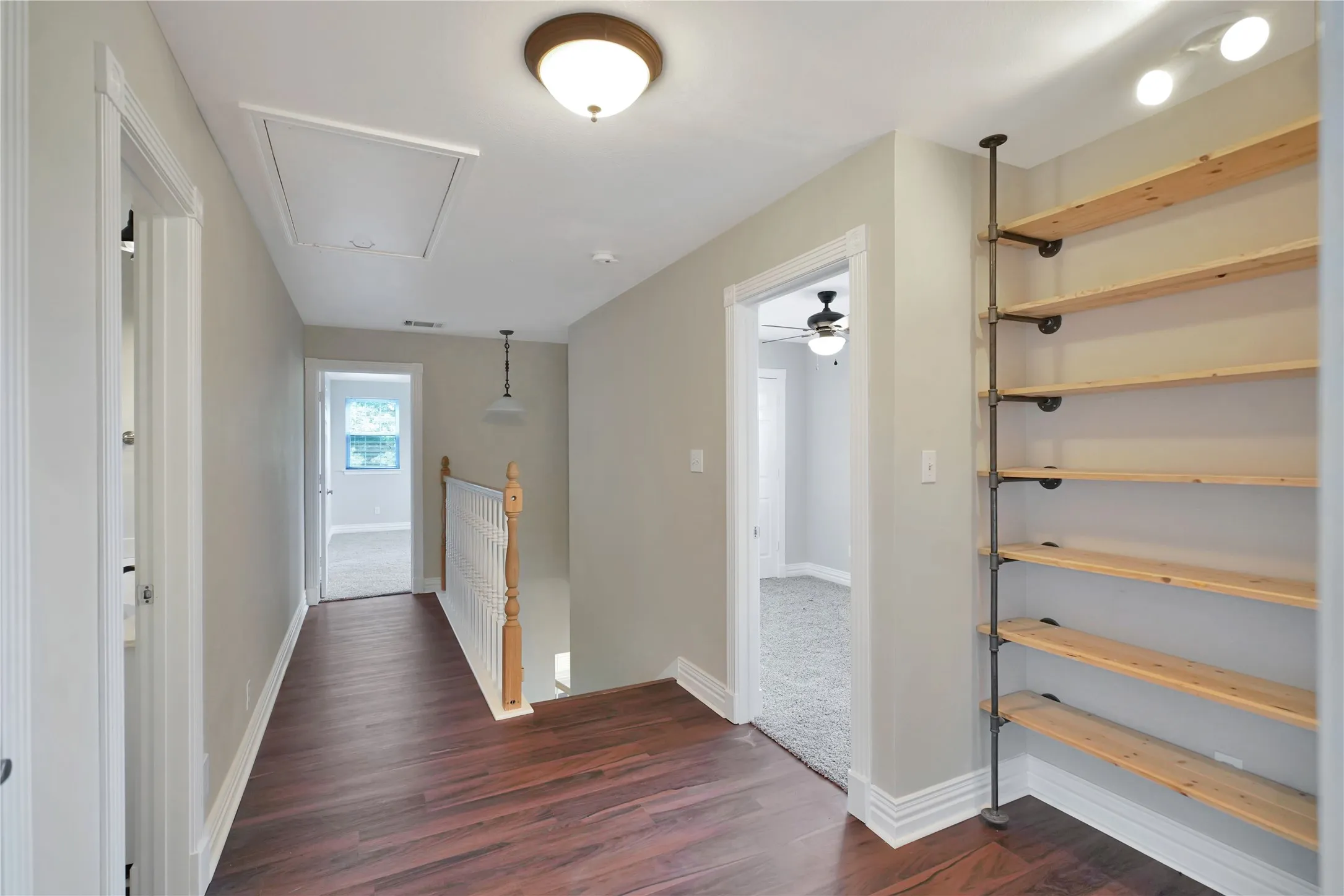 Wood-finish flooring, built-in open shelving, staircase with wood newel post and white balusters, overhead flush mount lighting, and attic access panel