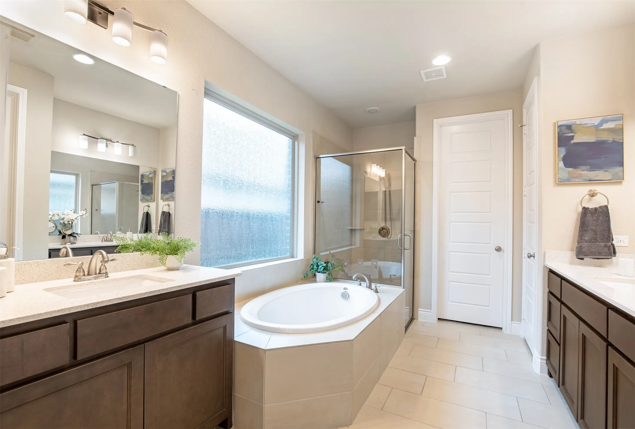 Dual vanities with undermount sinks, a soaking tub, a glass-enclosed shower, and light-toned floor tiling