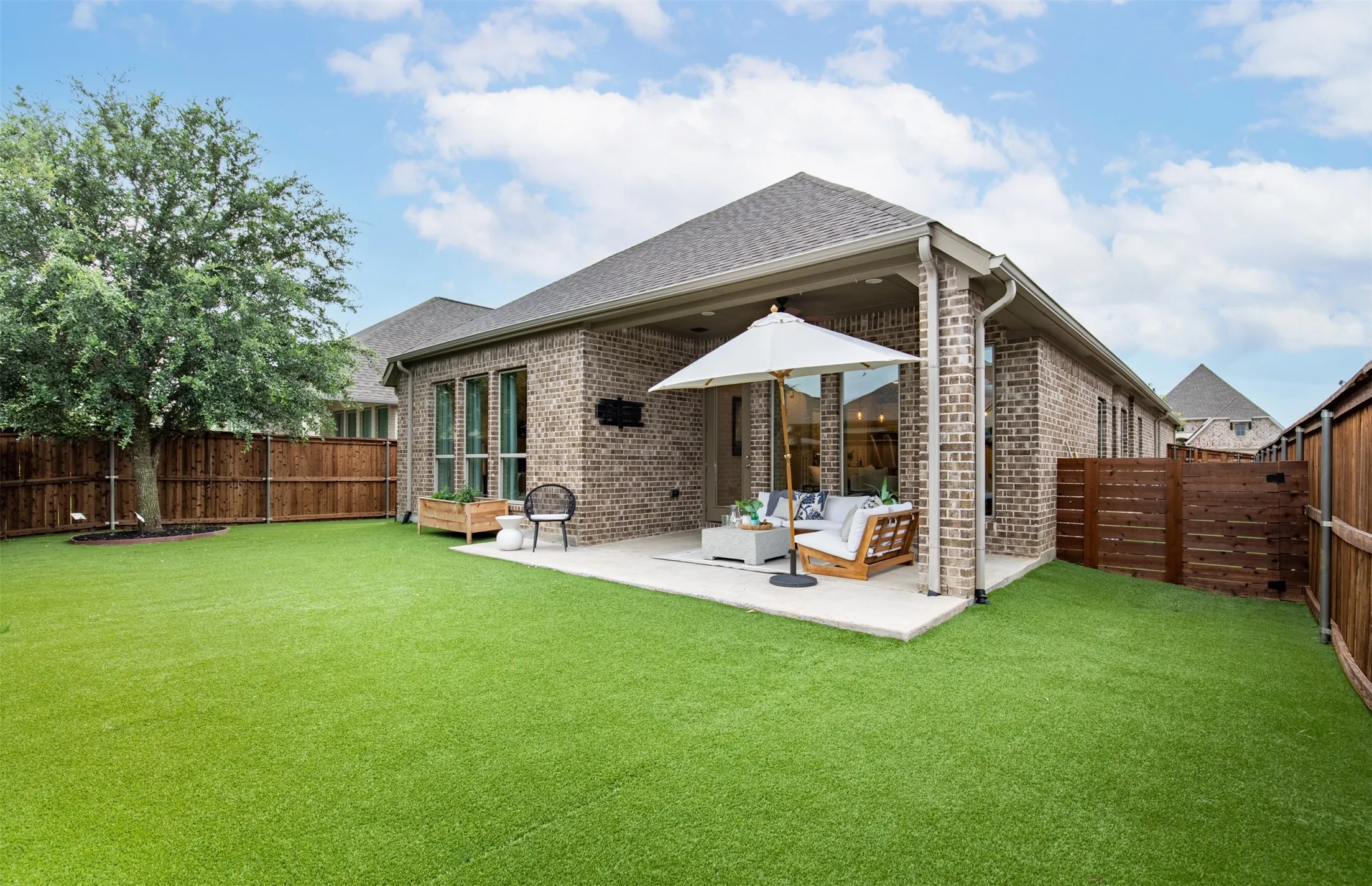 Brick exterior with covered patio, synthetic turf lawn, mature tree, and wood fencing