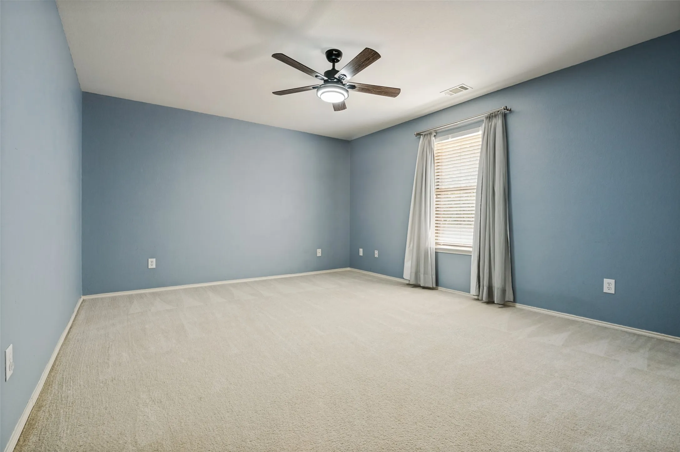 Expansive room featuring light-colored carpeting, blue walls, a window with blinds, and a ceiling fan with integrated lighting