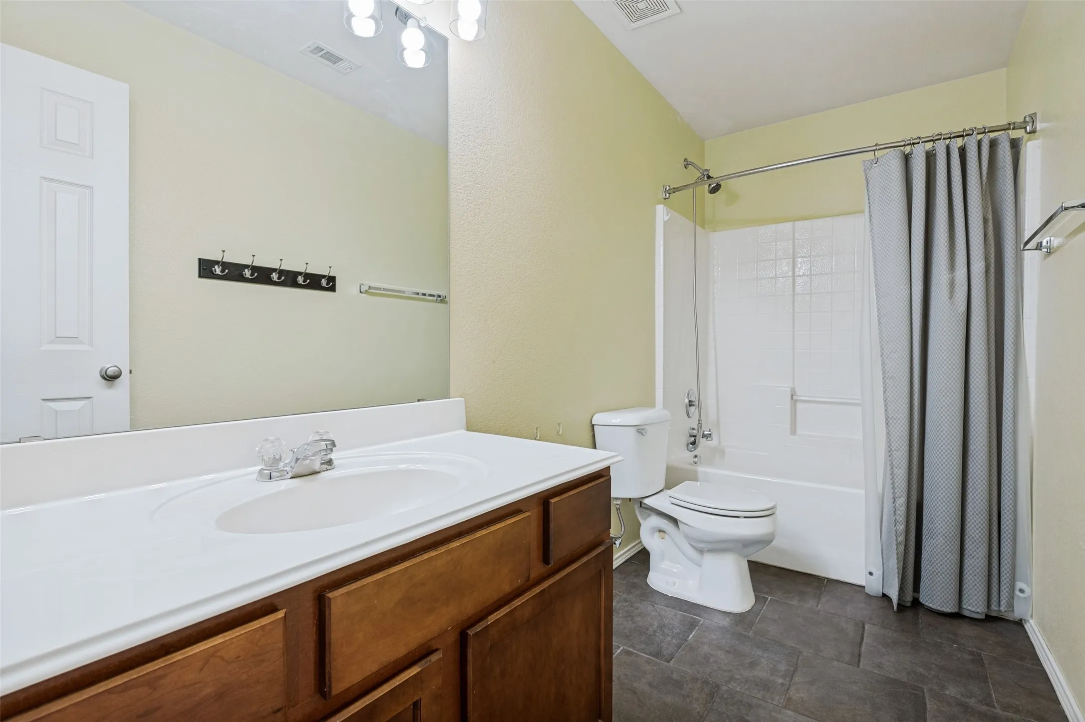 White bathtub with a shower, dark gray floor tiles, wood-finish vanity with an integrated sink, and an expansive wall mirror
