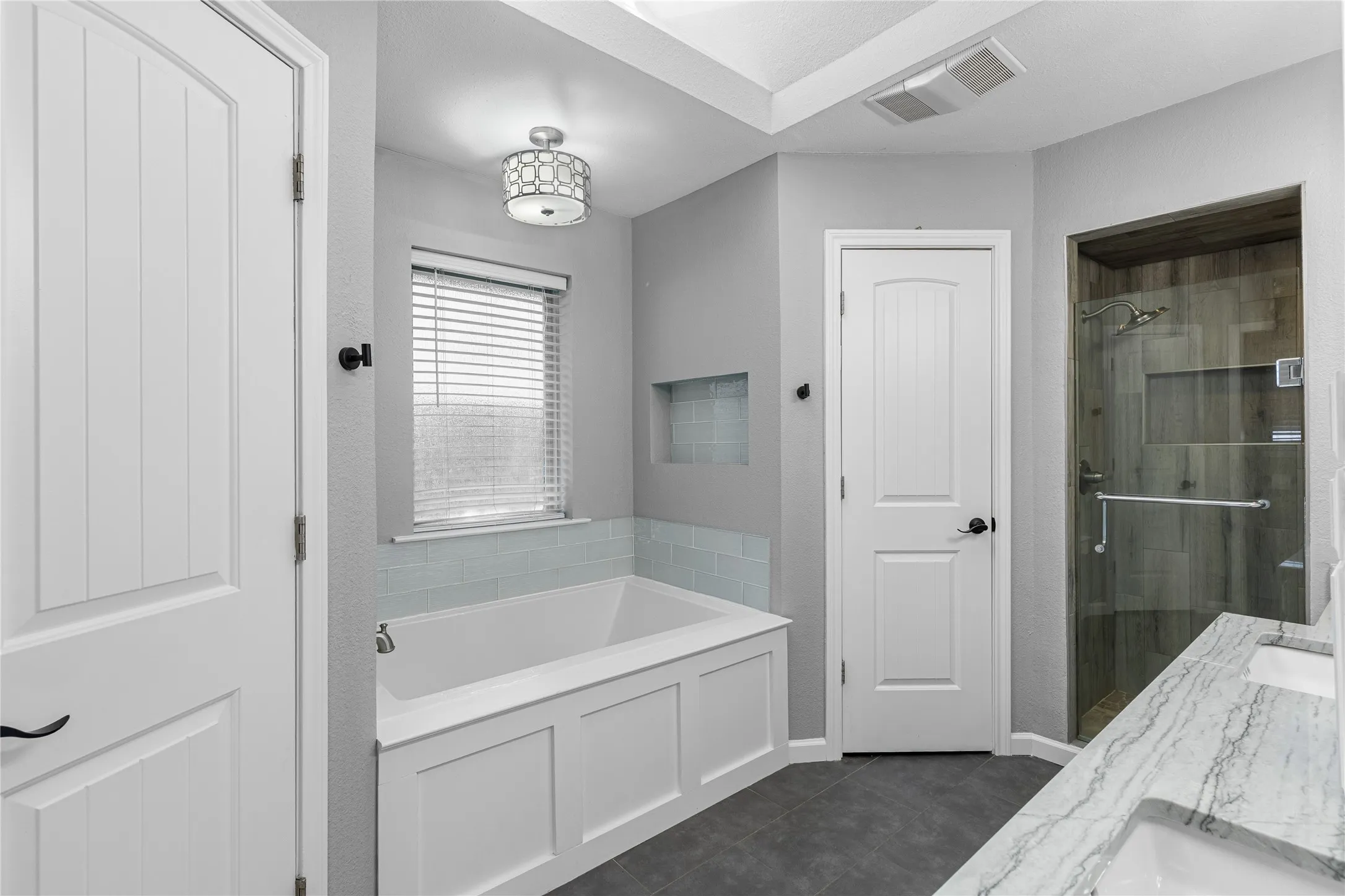 Soaking tub with subway tile surround, enclosed shower with wood-finish tile, dual vanity with undermount sinks, and grey tile flooring