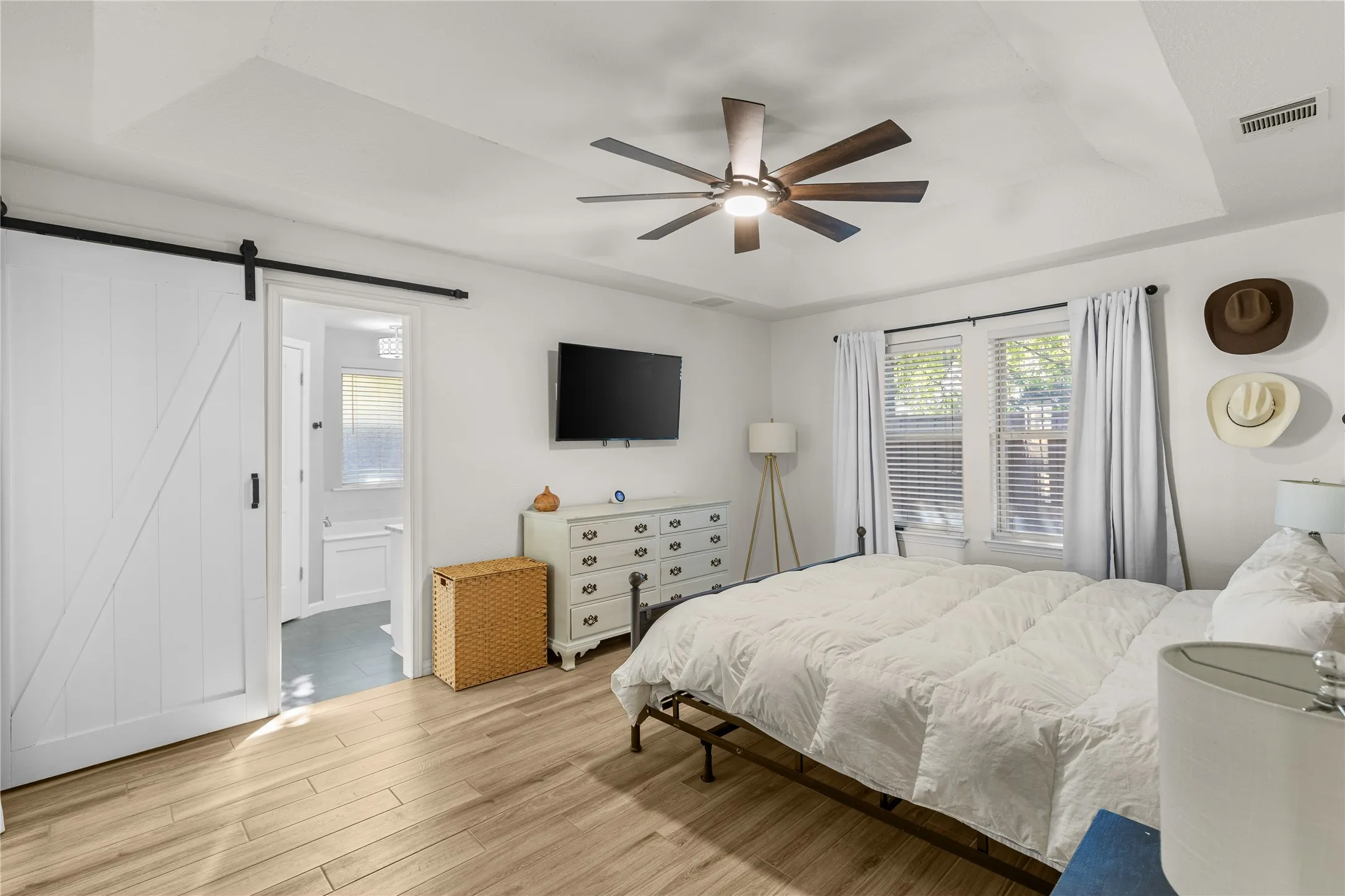 Wood-finish flooring, tray ceiling, modern ceiling fan, white barn door, and dual windows with blinds