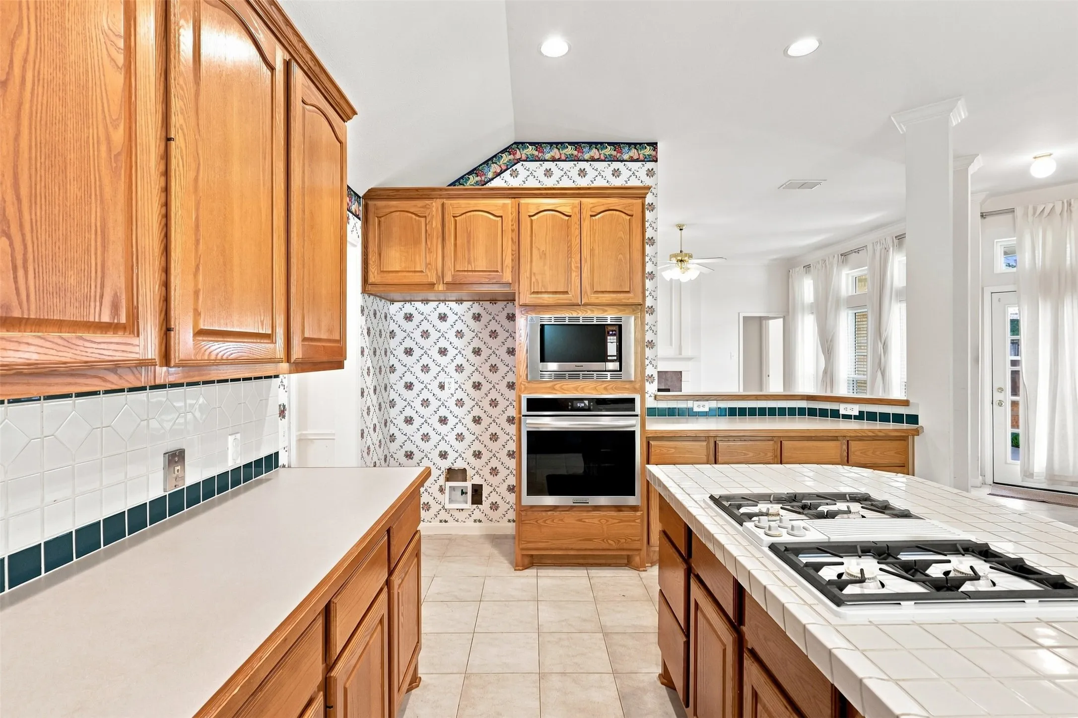 Wood cabinetry with stainless steel built-in appliances, tile backsplash, and a tile-top island with a cooktop.