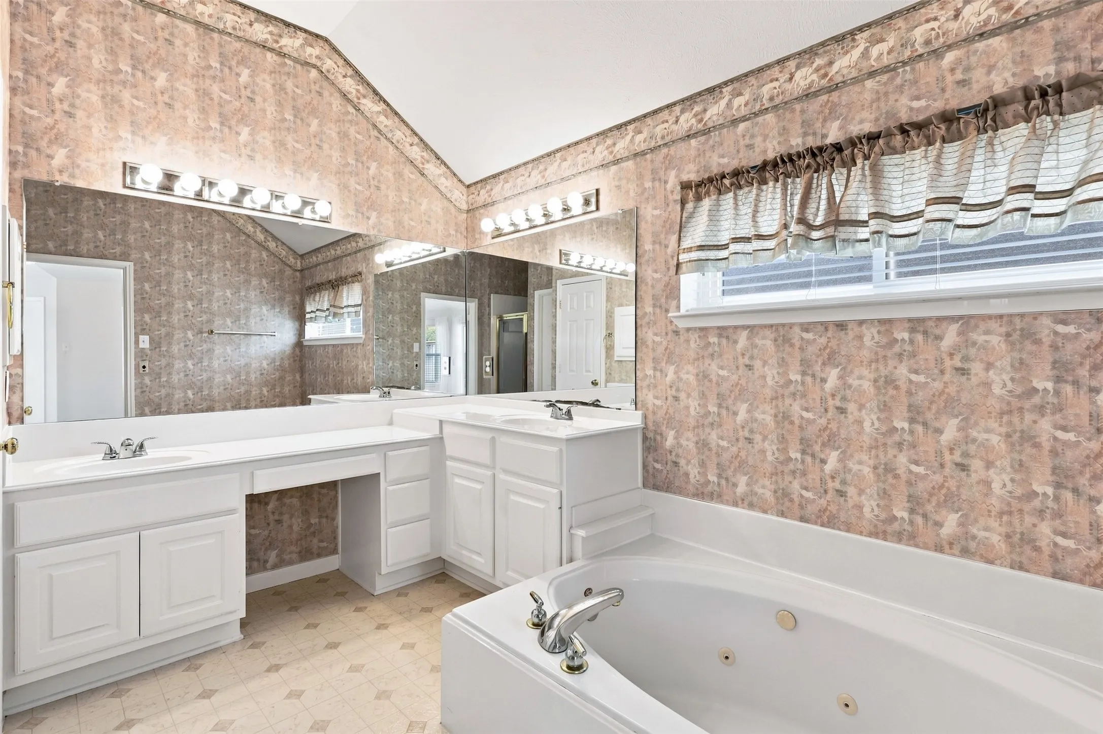 Primary bathroom features dual vanity sinks with white cabinetry, mirrored wall, recessed jetted bathtub, and patterned wallpaper.