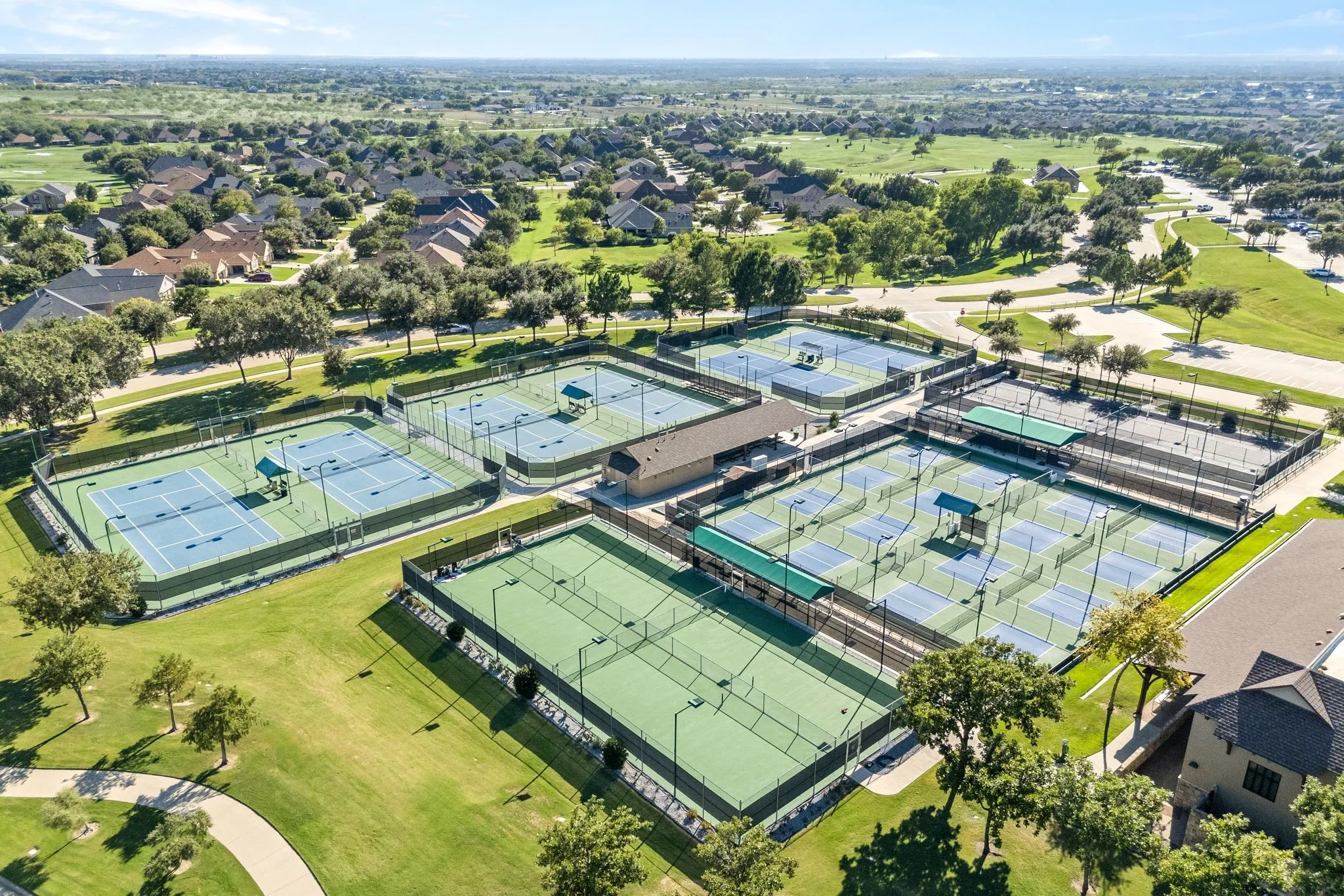 Multiple tennis and pickleball courts with perimeter fencing, shaded seating areas, and adjacent green spaces
