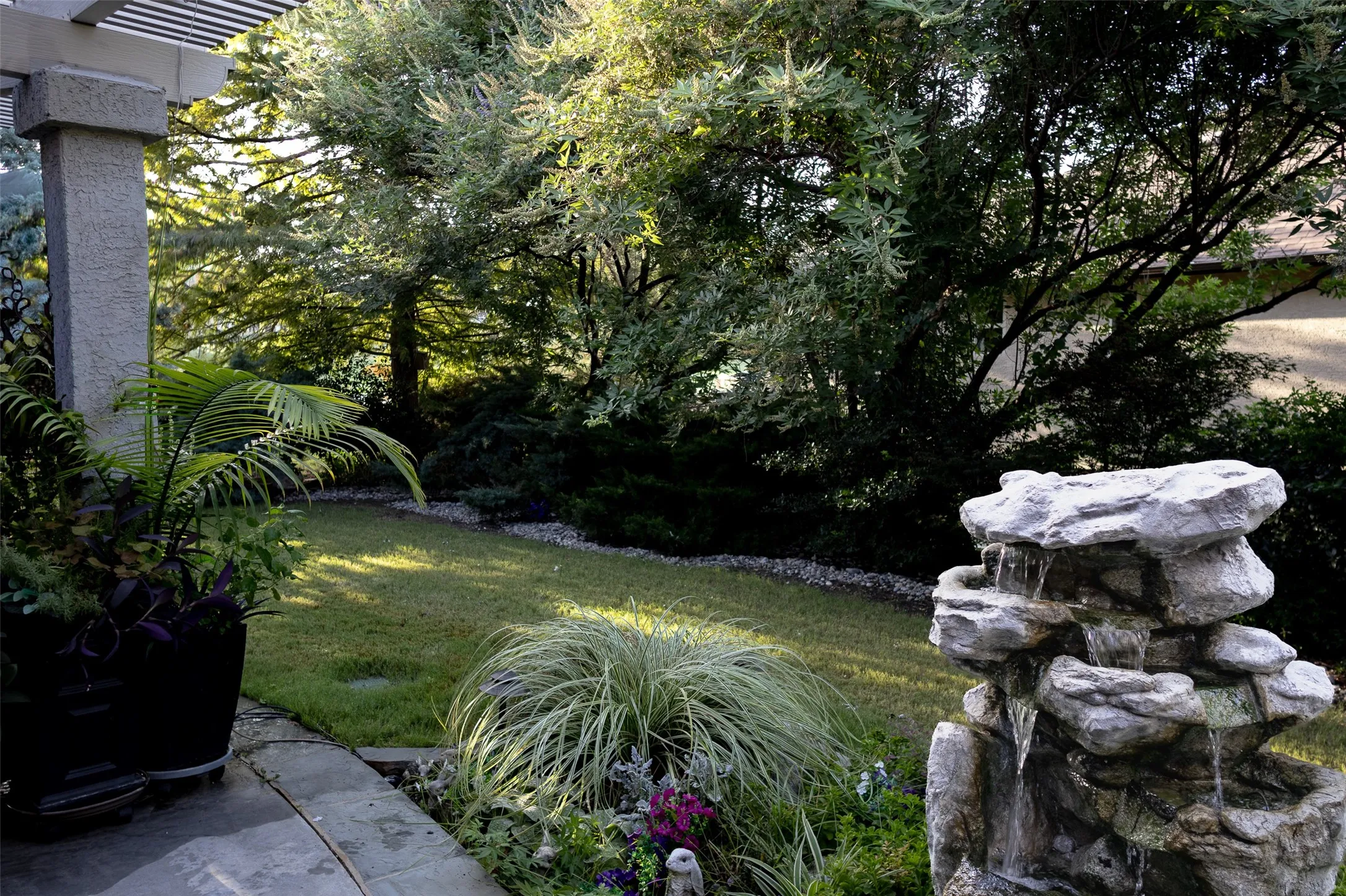 Stone patio with a slatted pergola, textured structural pillar, manicured lawn, and a multi-tiered rock water feature