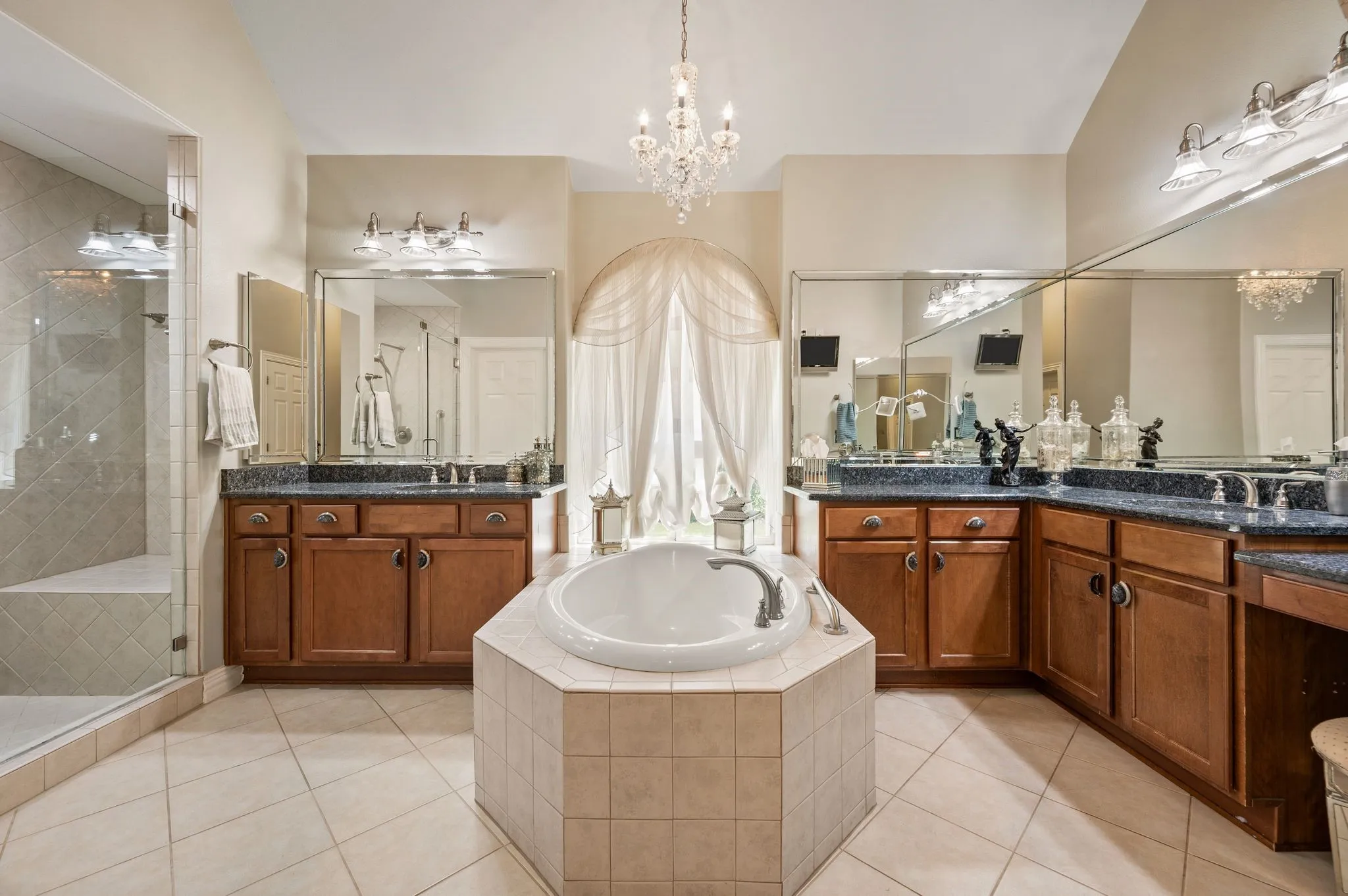 Built-in soaking tub, dual wood-finish vanities with dark countertops, tiled flooring, glass-enclosed shower, and a crystal chandelier