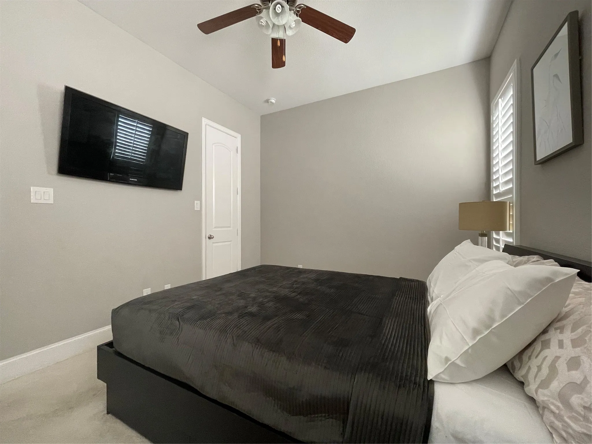 Neutral wall paint, ceiling fan, white door, window with plantation shutters, wall-mounted television