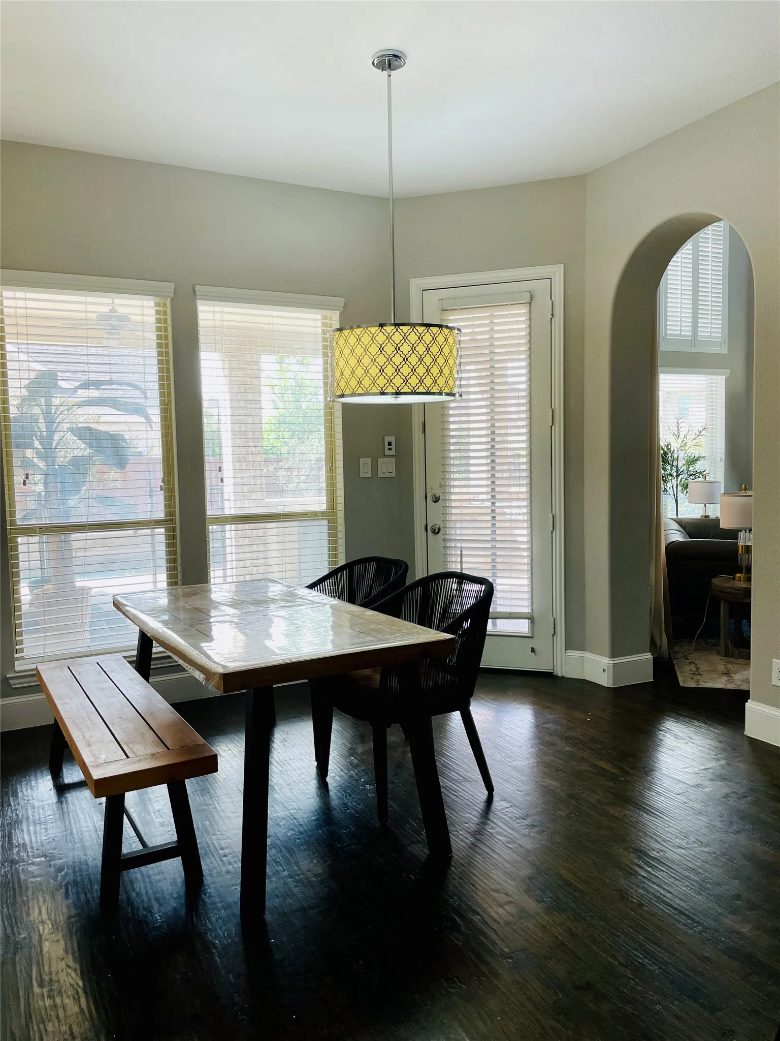 Dark wood-finish flooring, multiple windows with blinds, built-in archway, pendant light fixture, and a white door with blinds