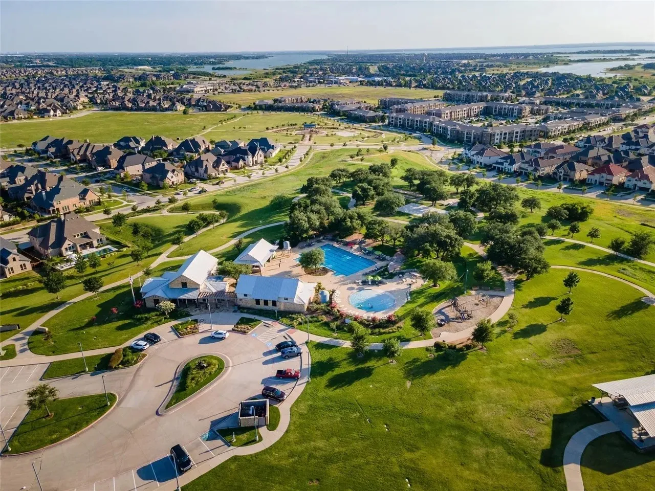 Community green space featuring a large central building with a metal roof, two swimming pools, paved pathways, and mature trees