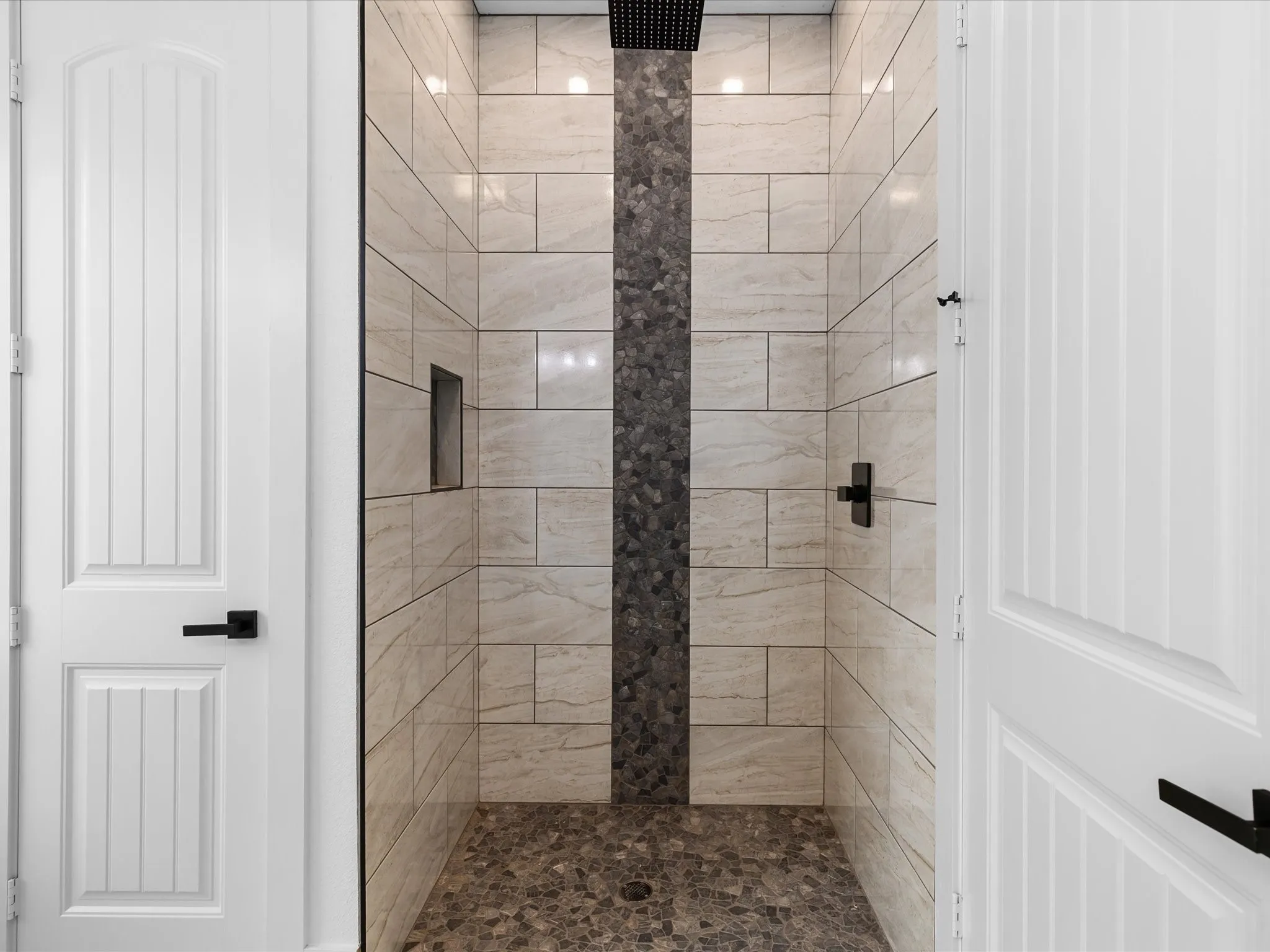 Walk-in shower featuring light-toned rectangular tile, a vertical accent strip of dark stone mosaic, a coordinating stone mosaic shower floor, an integrated shower niche, and matte black fixtures