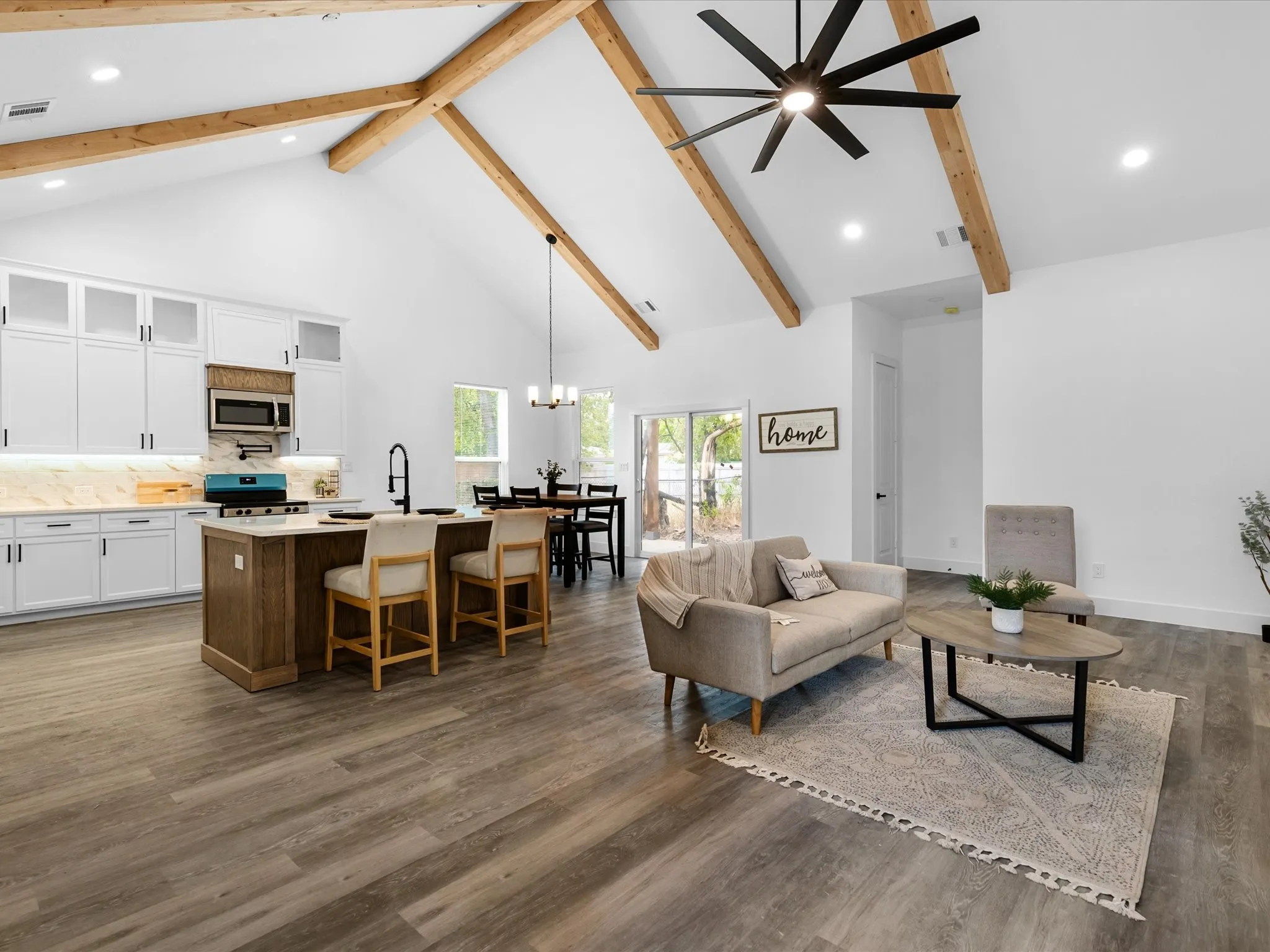 Vaulted ceiling with exposed wood beams, wood-finish flooring, white cabinetry with upper glass-front panels, kitchen island with seating, and a large ceiling fan