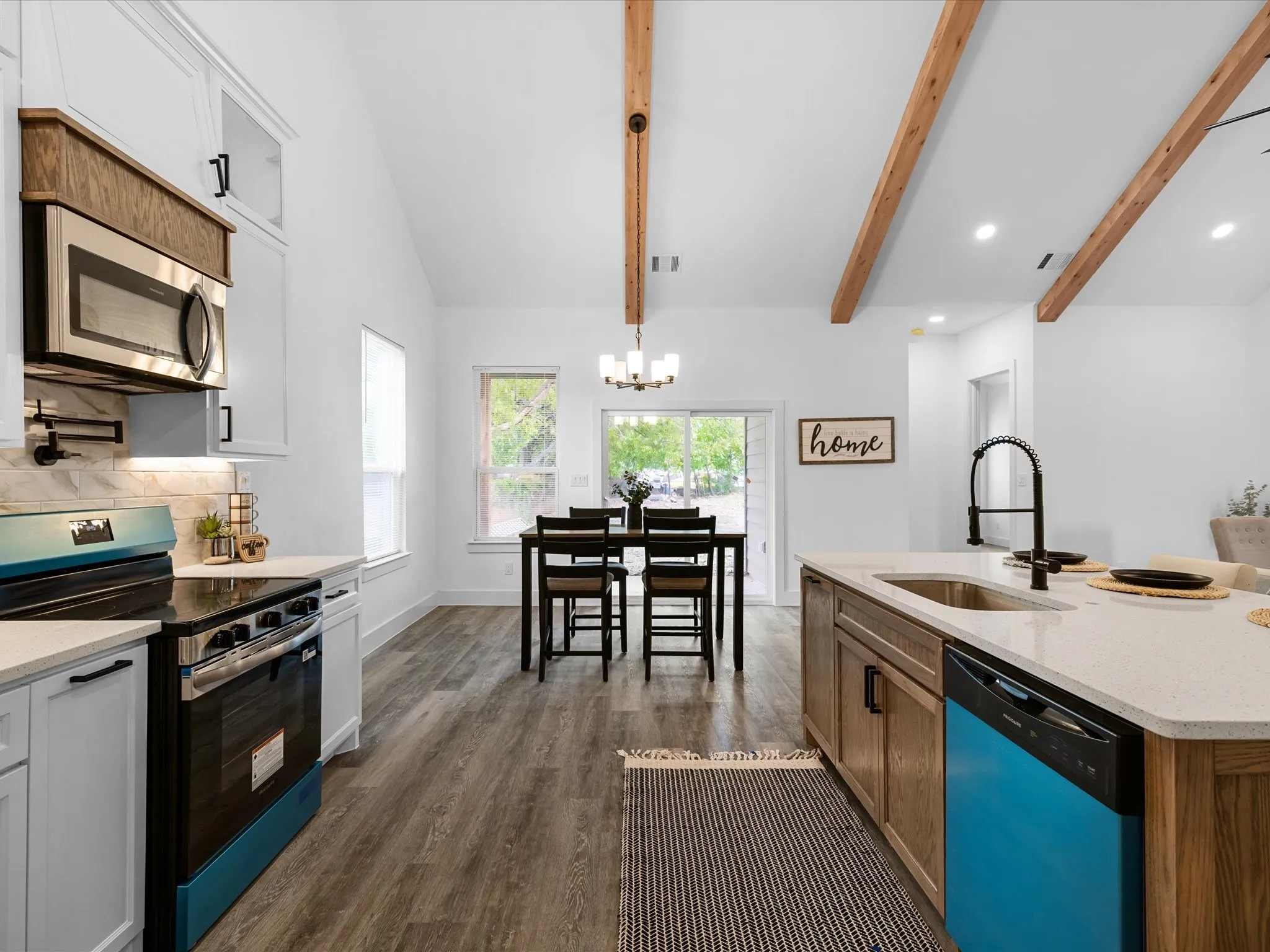 Vaulted ceiling with exposed wood beams, wood-finish flooring, white cabinetry, island with countertop and undermount sink, and stainless steel appliances