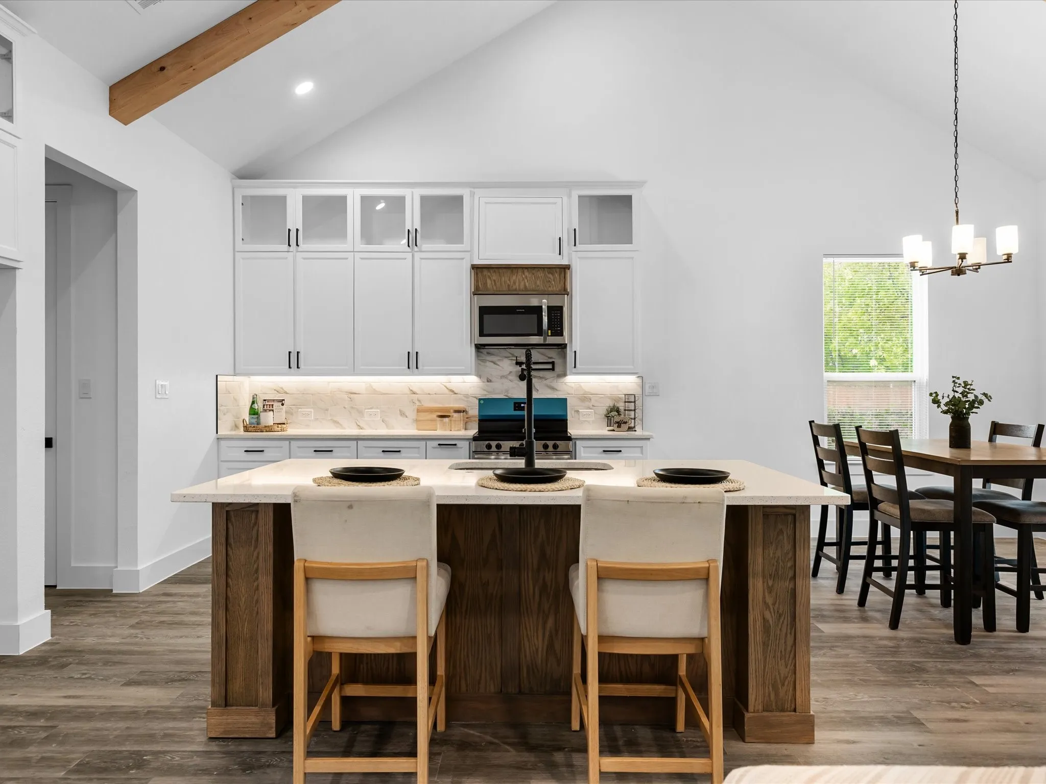 Vaulted ceiling with an exposed wood beam, white cabinetry with glass upper doors, under-cabinet lighting, kitchen island with a light-toned countertop, and wood-finish flooring