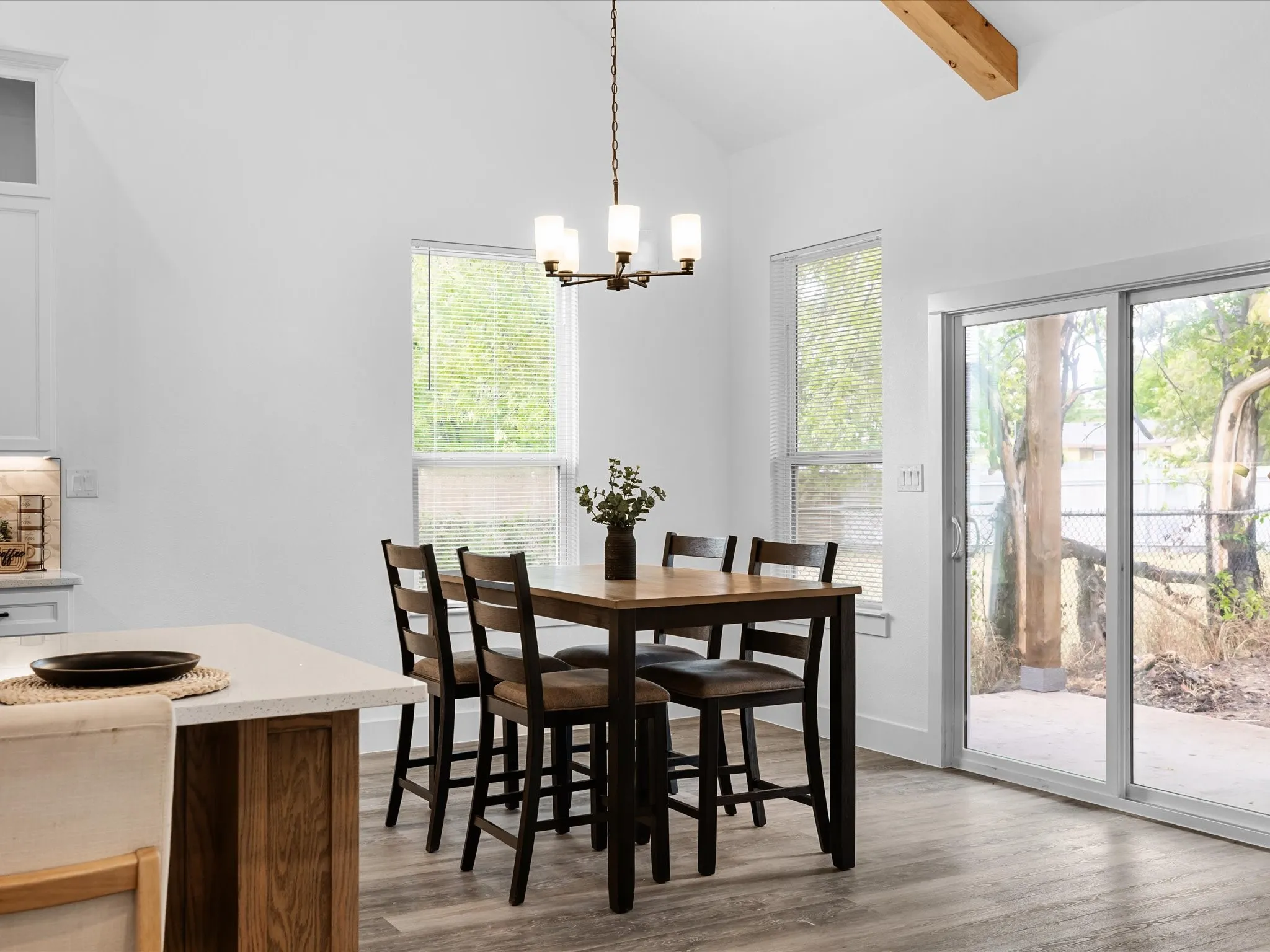 Vaulted ceiling with exposed wood beam, modern chandelier, wood-finish flooring, two large windows, and a sliding glass door
