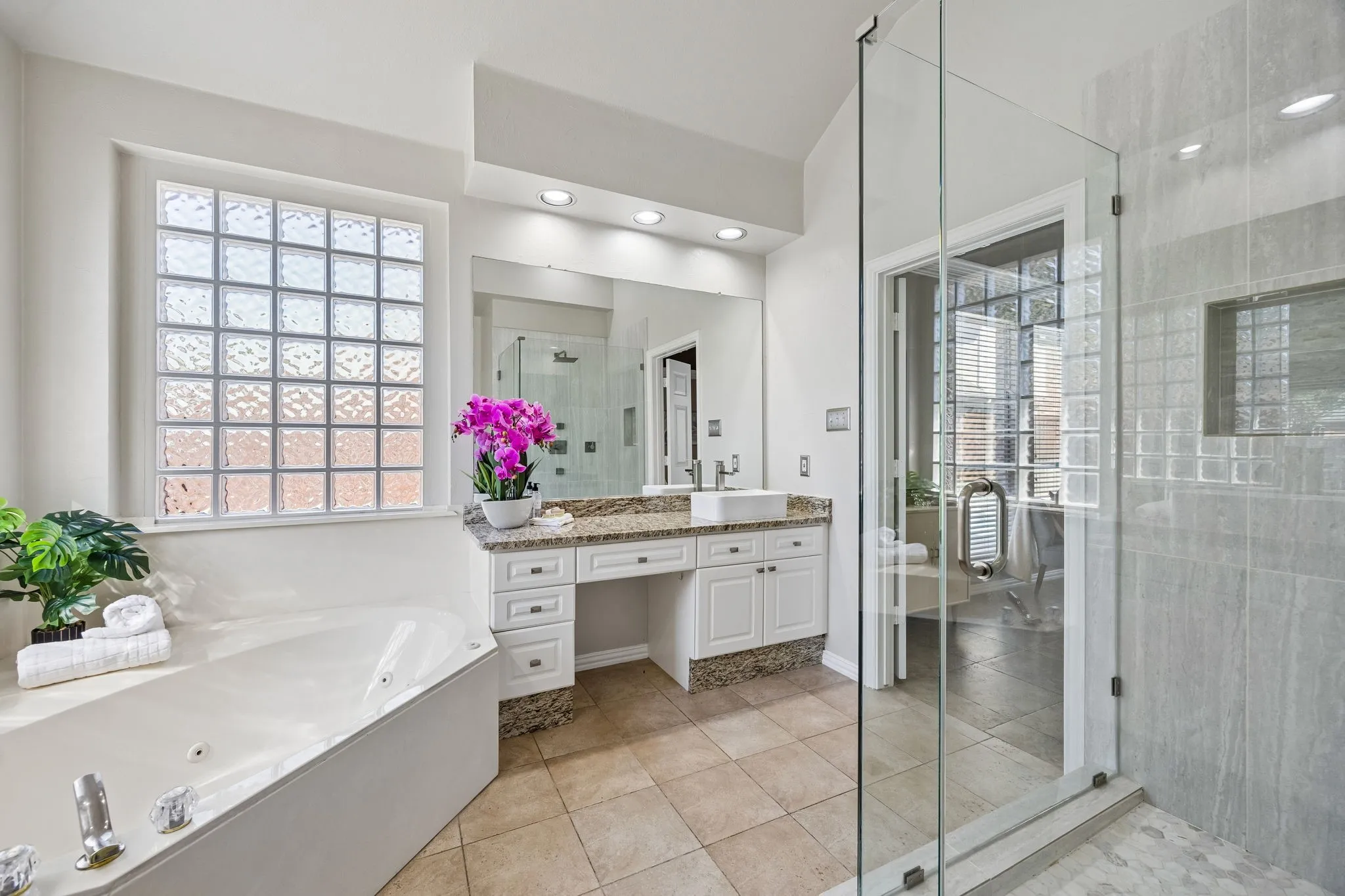 Spacious bathroom with a walk-in shower, soaking tub, glass block window, and dual vanities with stone countertops