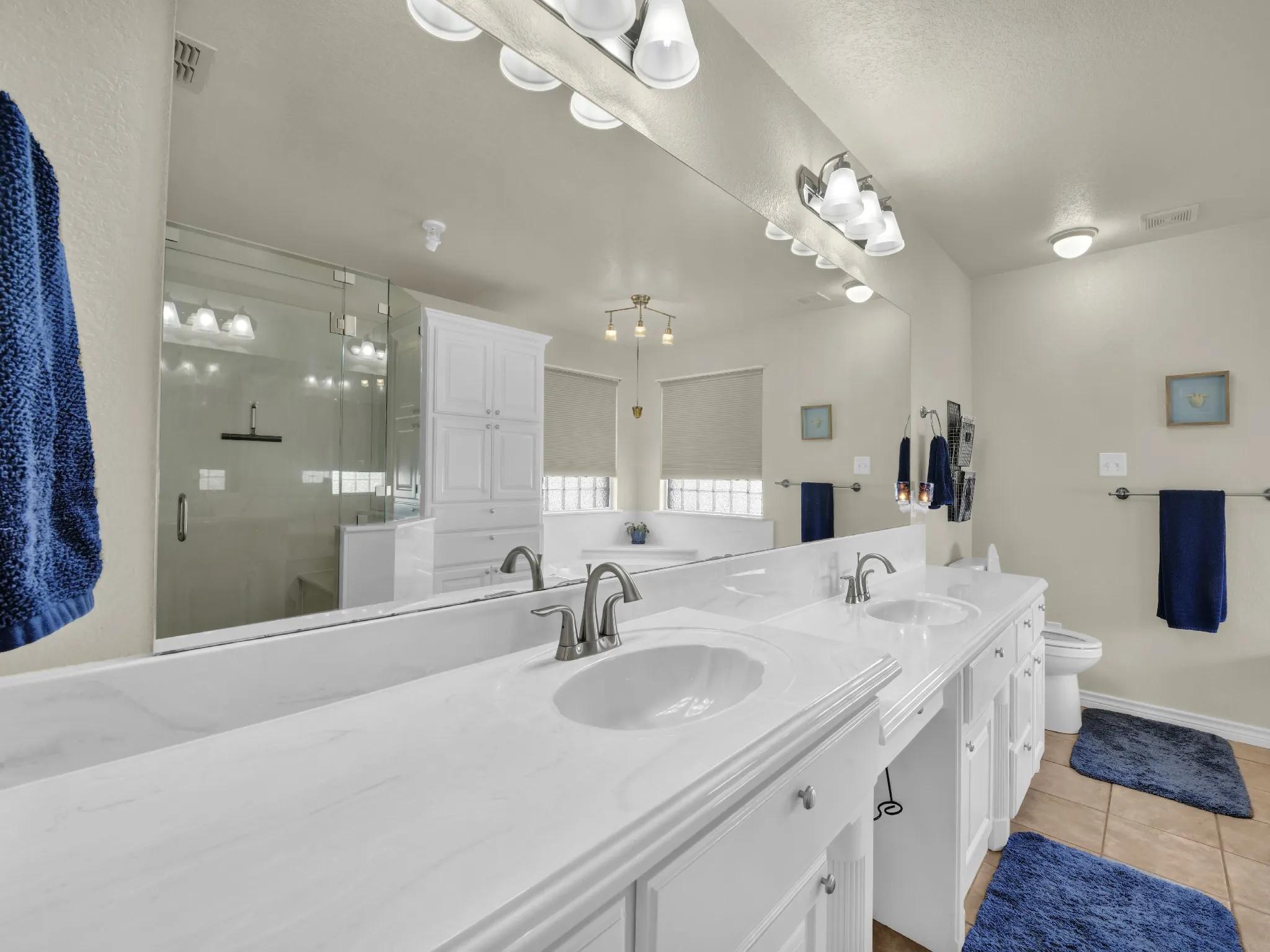 Dual vanities with undermount sinks, expansive mirror, glass-enclosed shower, white cabinetry, and tiled flooring