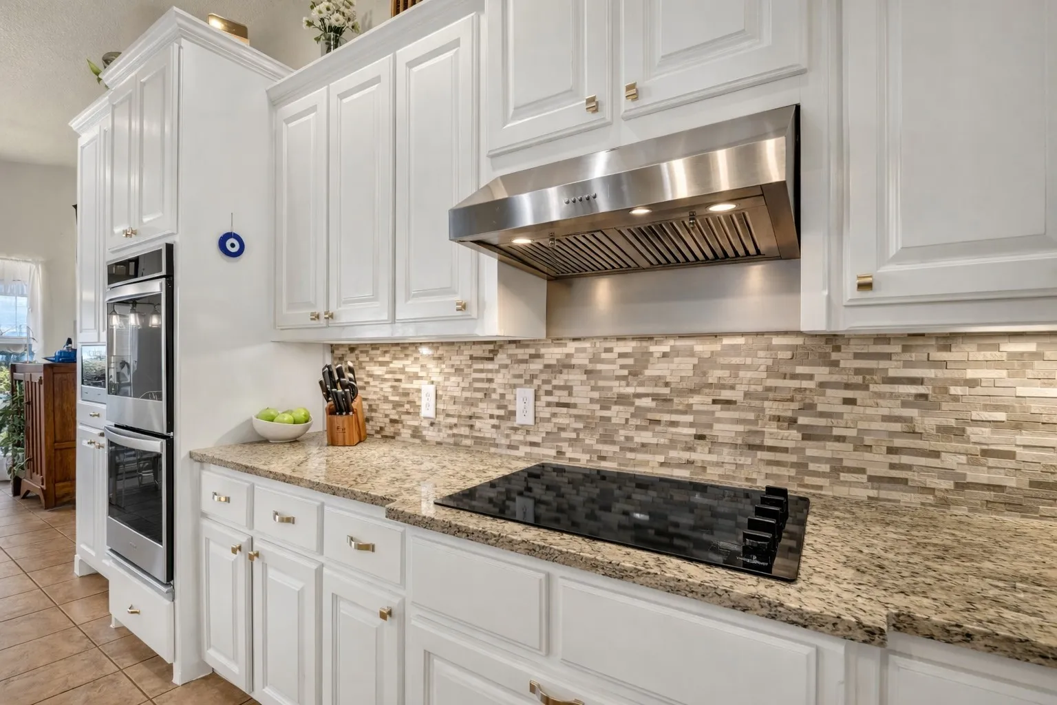 White cabinetry with gold-tone hardware, a mosaic tile backsplash, a built-in cooktop, and stainless steel double ovens
