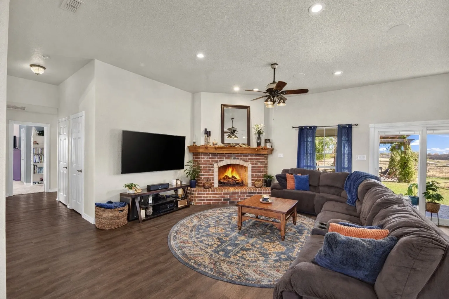 Corner brick fireplace with wood mantel, recessed lighting, ceiling fan, dark wood-finish flooring, and sliding glass door