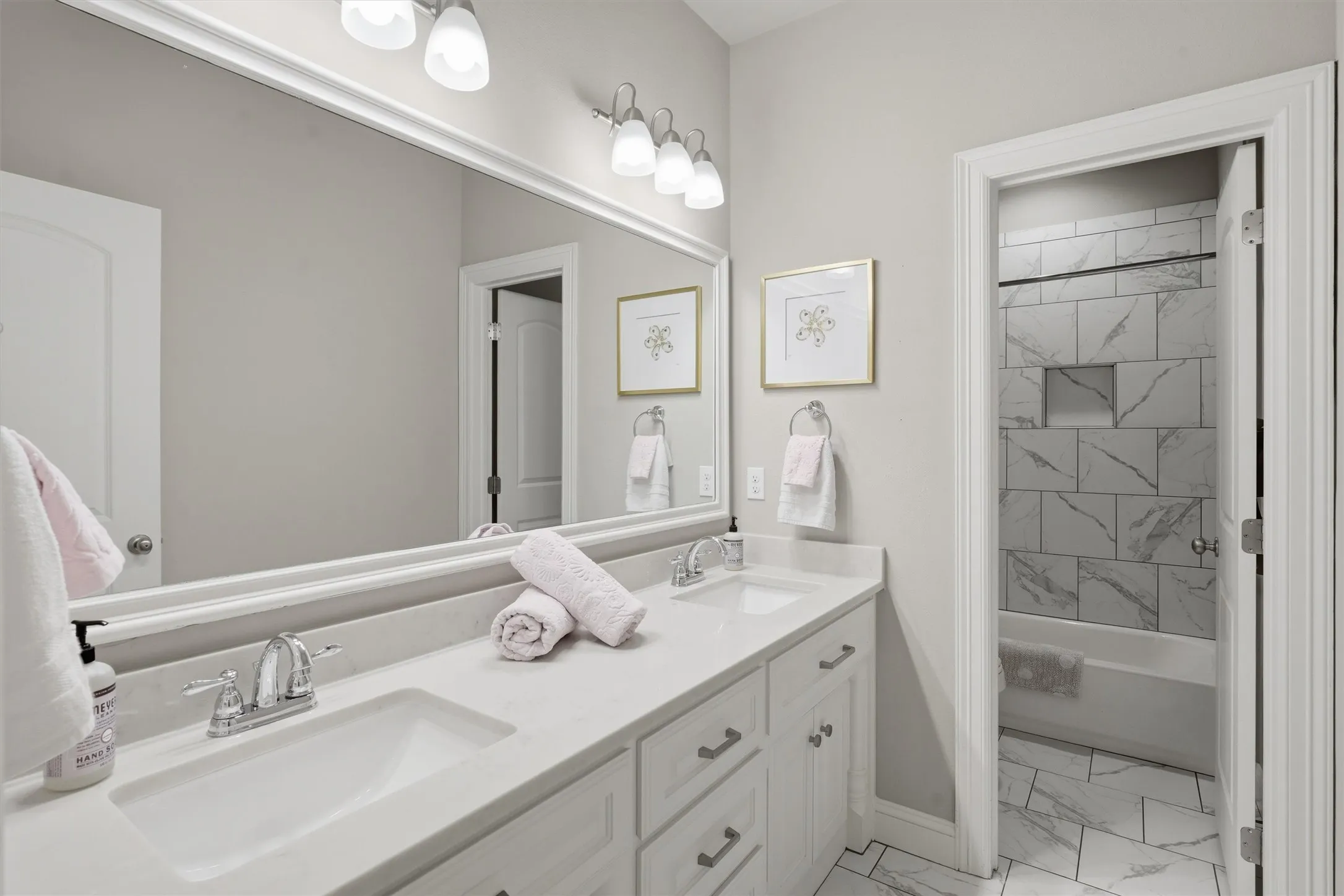 This well-appointed secondary bathroom features a double-sink vanity with abundant counter space, an oversized mirror, and bright vanity lighting. A private adjacent door leads to the shower-tub combo detailed with stylish marble-look tile surround and built-in storage niche.