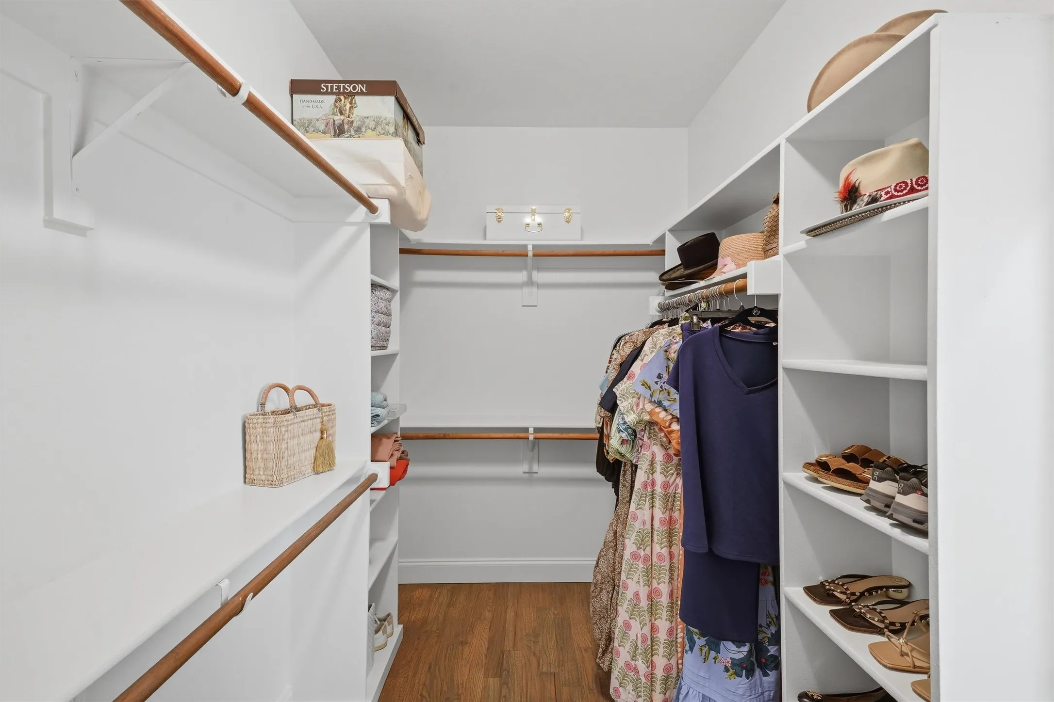 This spacious walk-in closet maximizes storage with custom built-in shelving, shoe displays, and double-tiered hanging rods. Warm wood flooring complements the clean, white custom organization system, making daily wardrobes easy to manage.