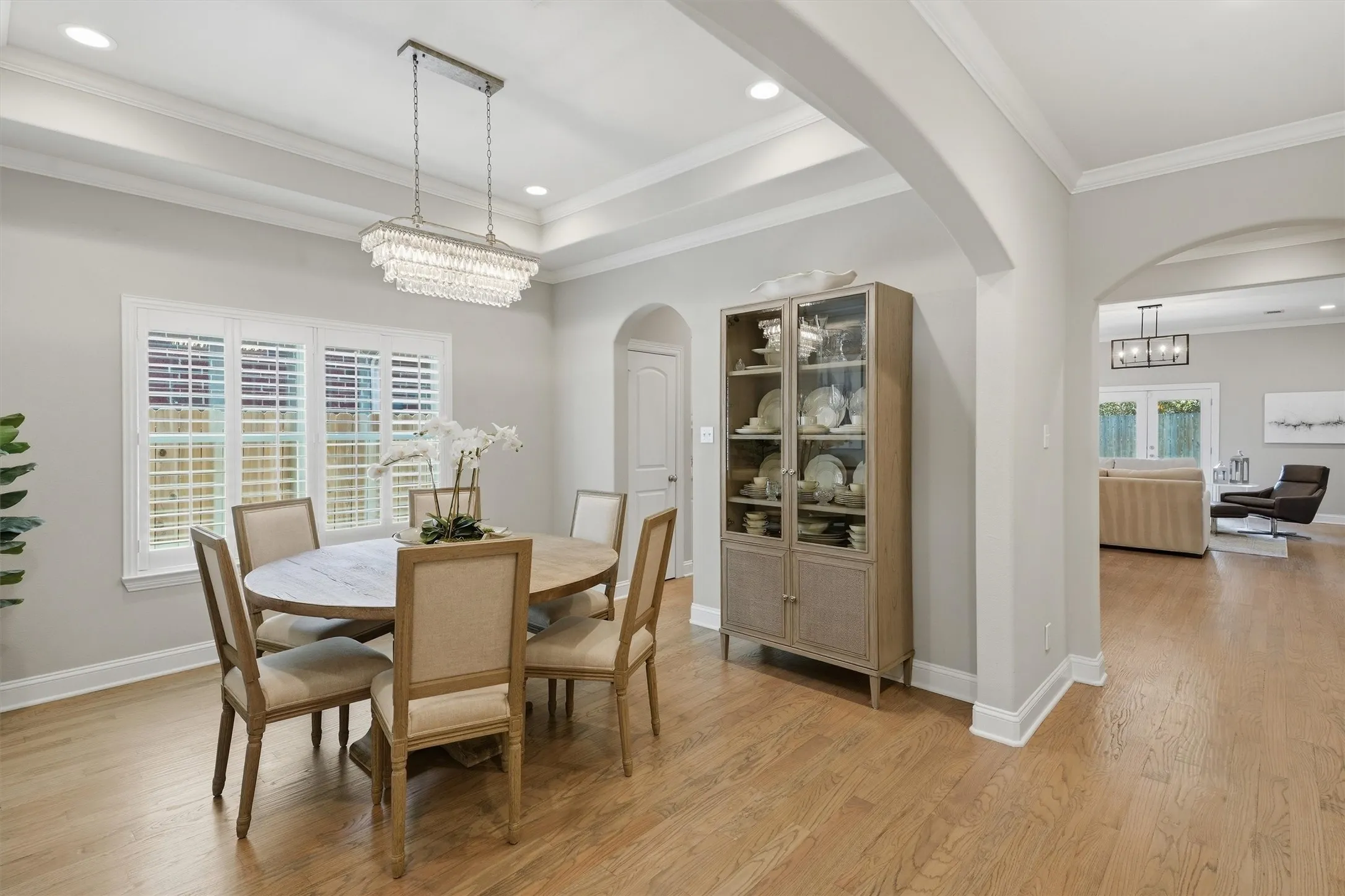 An elegant dining area highlighted by a recessed tray ceiling, glamorous crystal chandelier, and wide windows with plantation shutters. Graceful arched doorways frame views into the adjacent living space while maintaining a distinct, sophisticated setting for family meals.