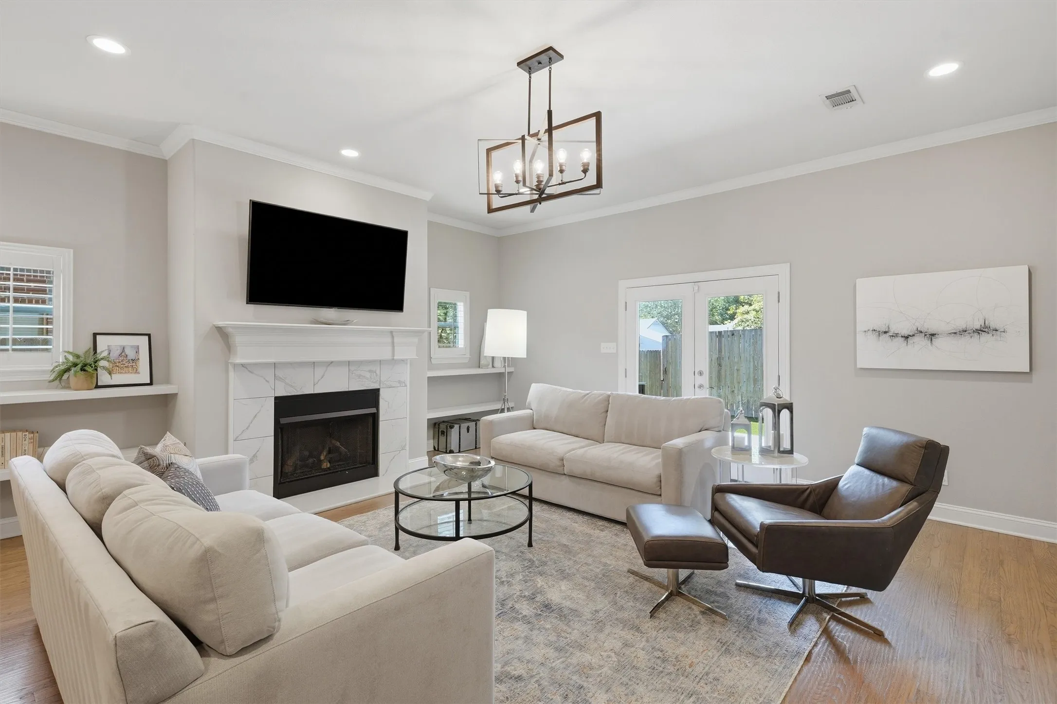 A focal marble-look tile fireplace anchors this bright living space, crowned by an elegant statement light fixture. Glass-panel French doors draw in natural light while providing direct access to the private backyard.