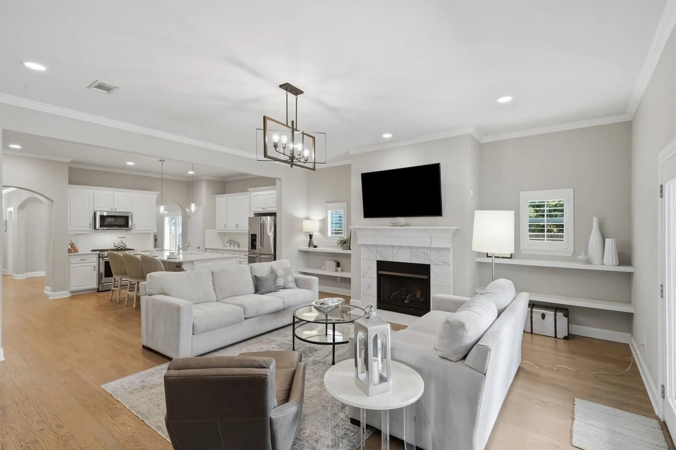 This spacious living room features a elegant tiled fireplace with a TV niche above, framed by sleek floating shelves and a statement chandelier. Large plantation-shuttered windows fill the open-concept space with soft light, seamlessly connecting the lounge area to the kitchen.