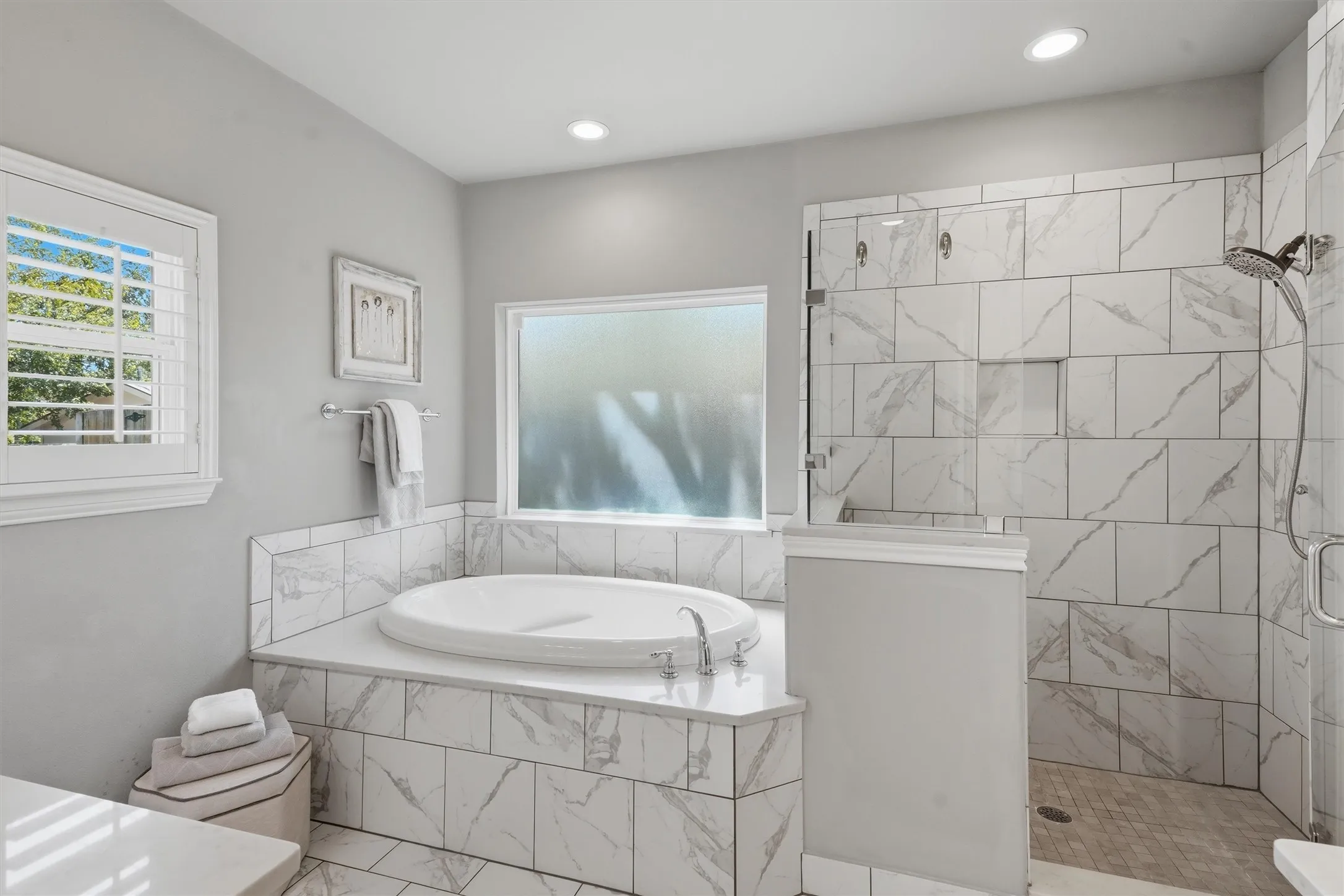 This spa-like primary bathroom showcases a deep oval soaking tub framed by elegant marble-look tile, paired with a seamless glass walk-in shower. Dual windows—including a privacy-frosted glass pane—fill the space with bright, calming light.