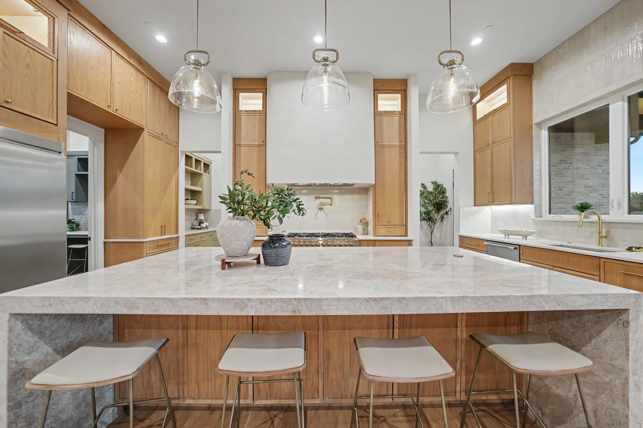 Expansive kitchen island with a light-toned countertop, clear pendant lighting, built-in cabinetry, stainless steel appliances, and a window with natural light