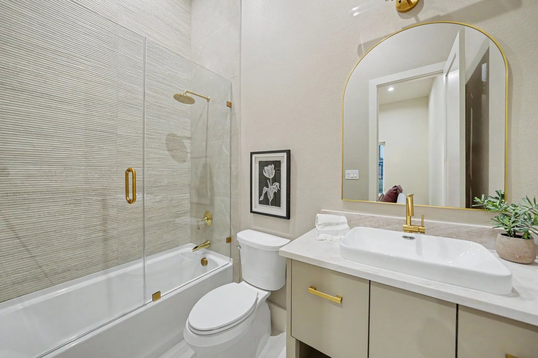 Bathroom featuring a tub with glass enclosure, textured tile surround, wall-mounted showerhead, single vanity with vessel sink, and arched mirror with gold-tone accents
