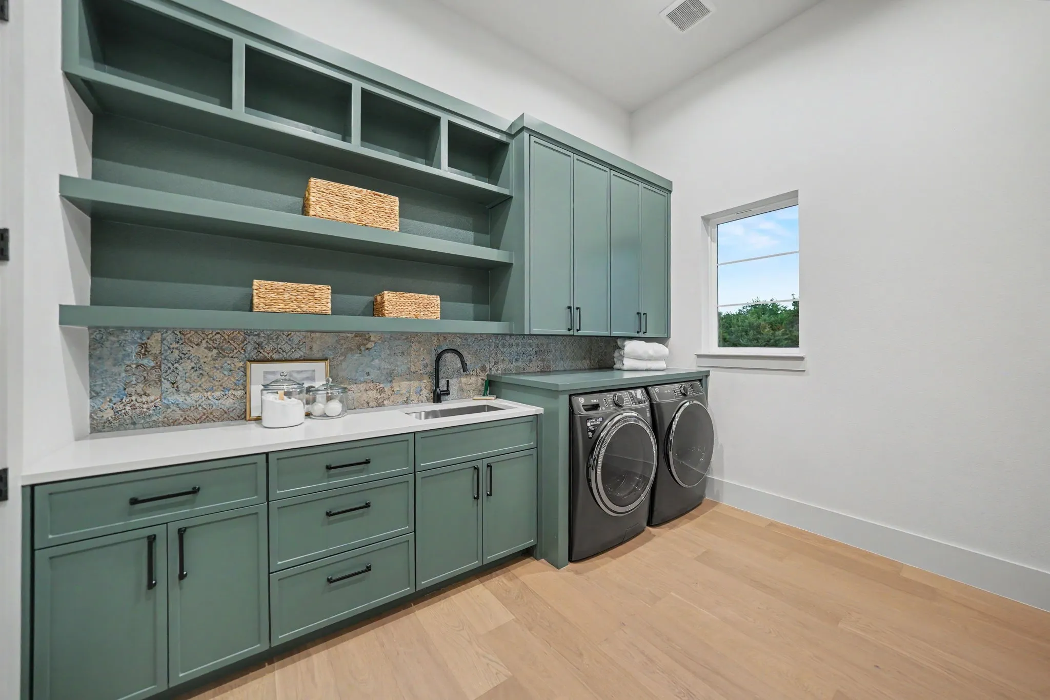 Custom cabinetry with integrated open shelving, quartz countertop, patterned tile backsplash, utility sink with a matte black faucet, and wood-finish flooring
