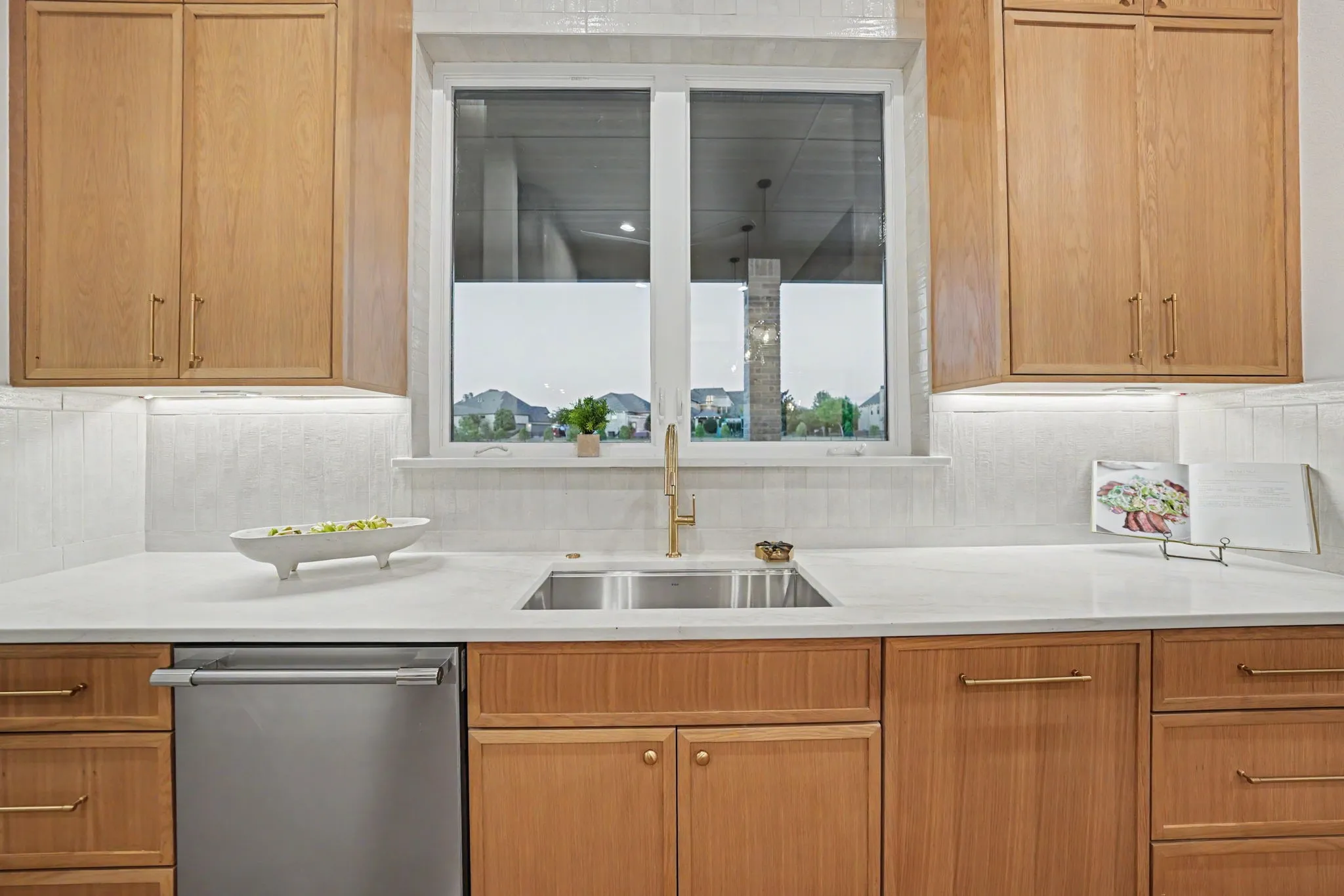 Light wood cabinetry with brass hardware, undermount double-basin sink with a gold-tone faucet, white solid surface countertops, and a full-height tile backsplash with under-cabinet lighting